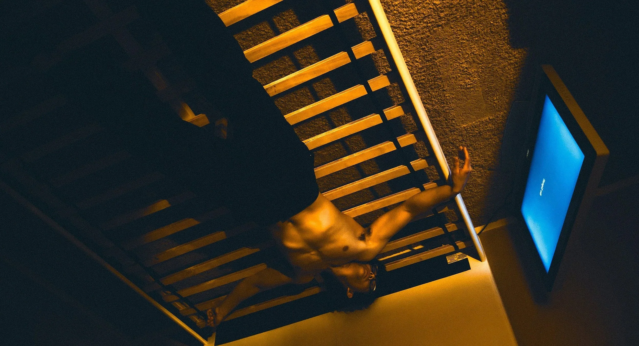 A person lying on their back on a wooden slat bed, reaching towards a blue digital device, in a dimly lit room.