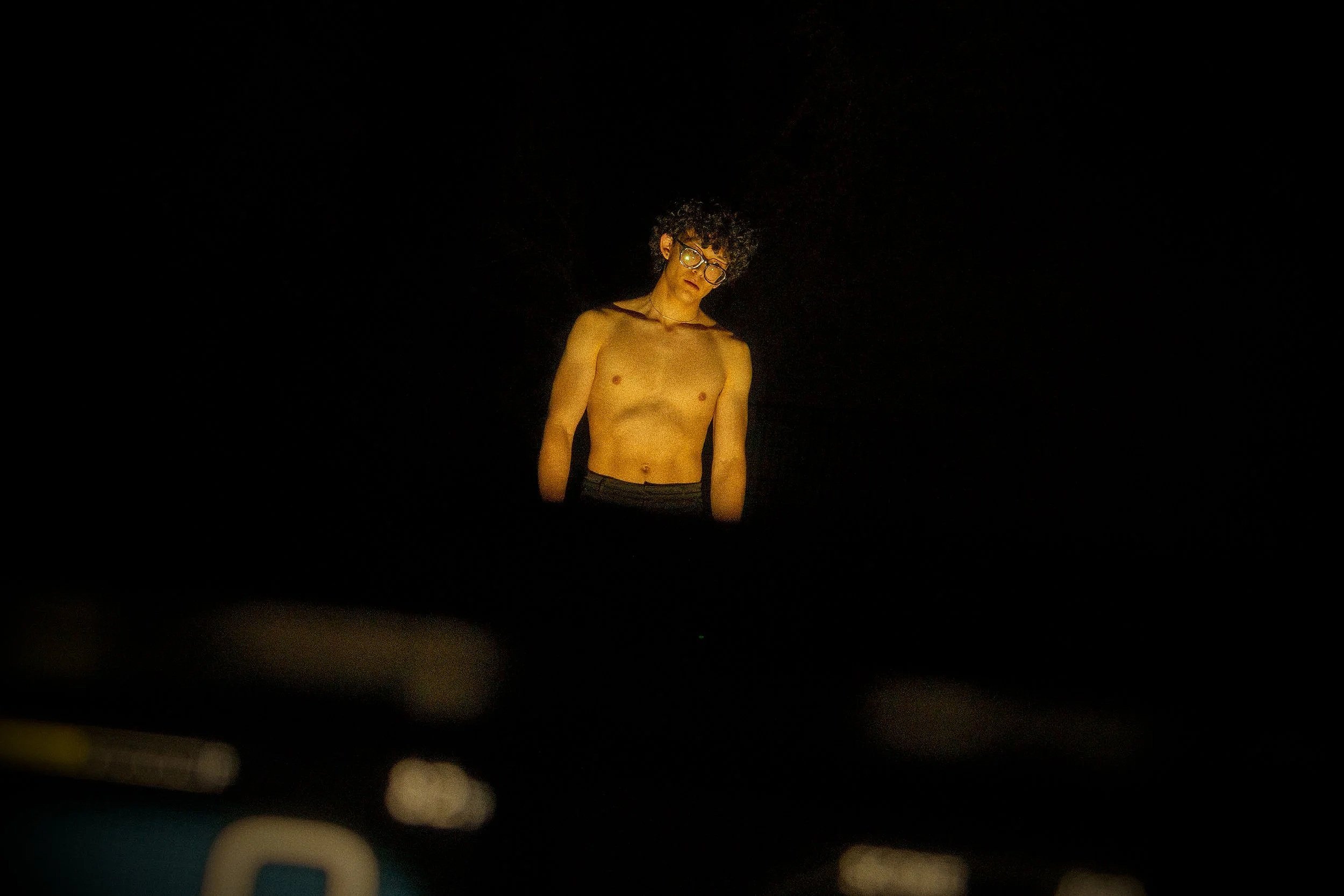 A shirtless person with glasses and curly hair standing in a dark room illuminated by a faint light.