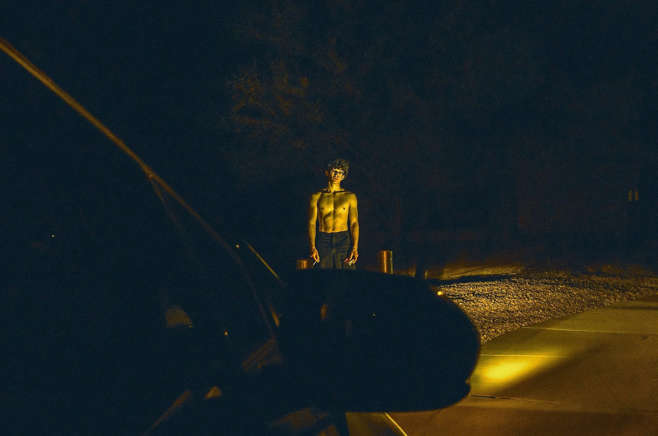 A shirtless man with glasses standing outdoors at night under a dark sky, illuminated by a nearby light source.
