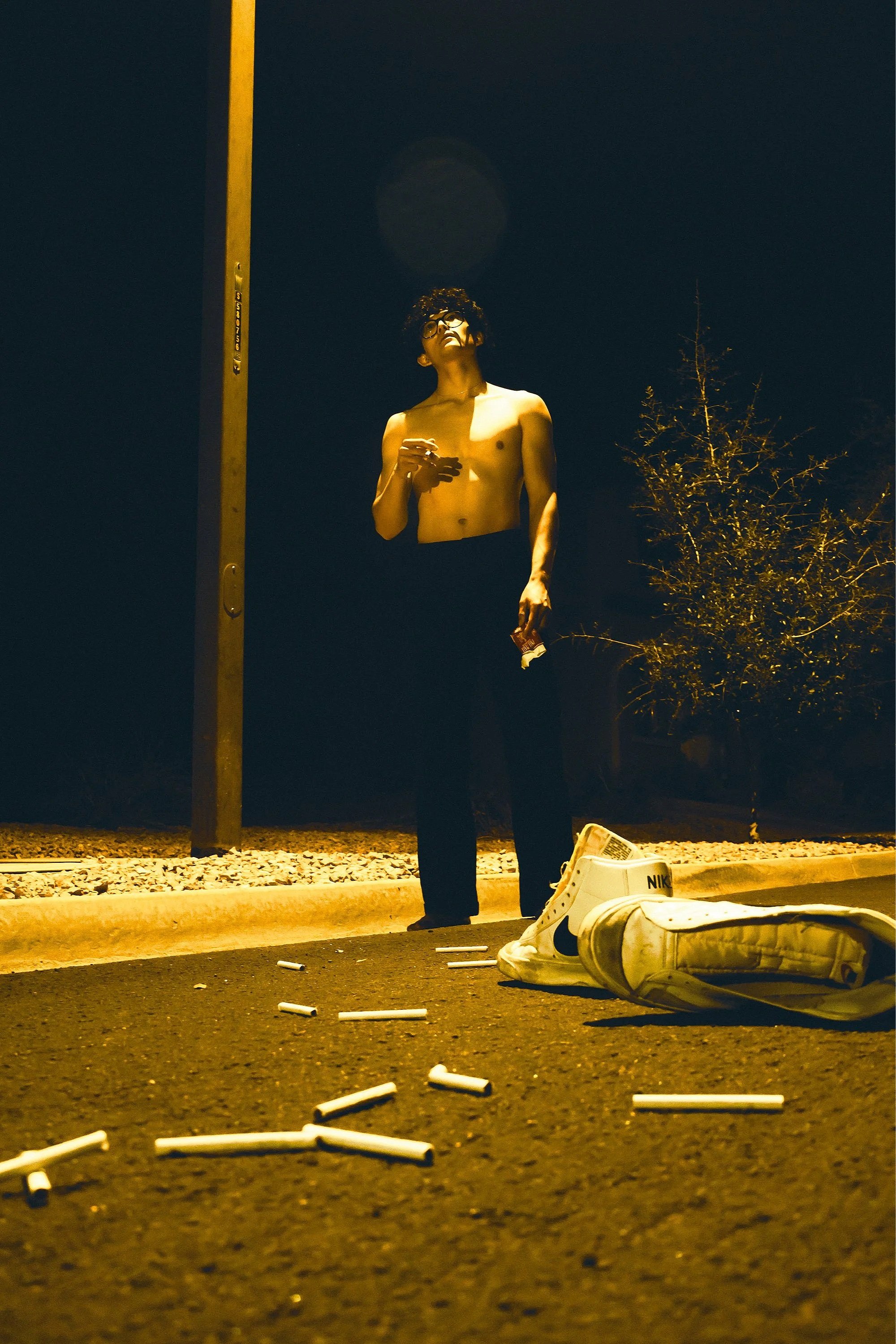 A shirtless young man with glasses stands on a street at night, holding a phone in one hand and a small object in the other. There are discarded cigarette butts on the ground, a white Nike sneaker with a black swoosh on the pavement, and a pair of wh