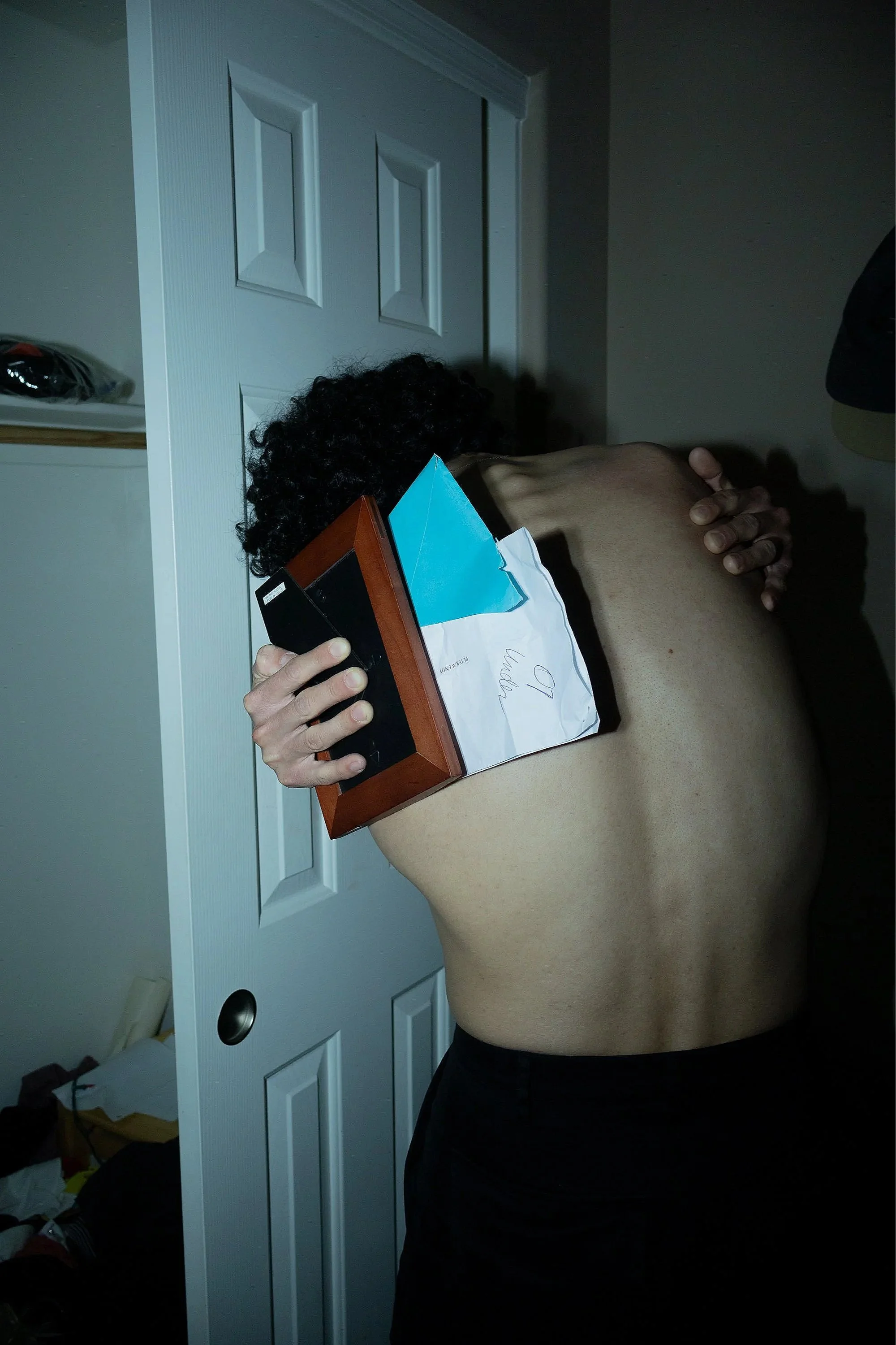 A person with curly dark hair holding a box with an envelope on top, standing in a room with white closet doors, wearing black pants, with their back and side visible.