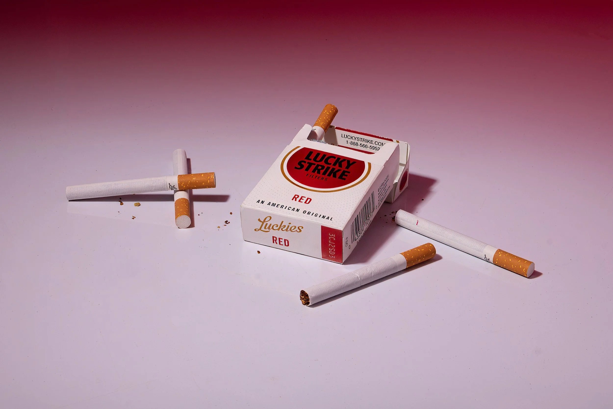 A pack of Lucky Strike cigarette packs with scattered cigarette butts and loose cigarettes on a pink surface.