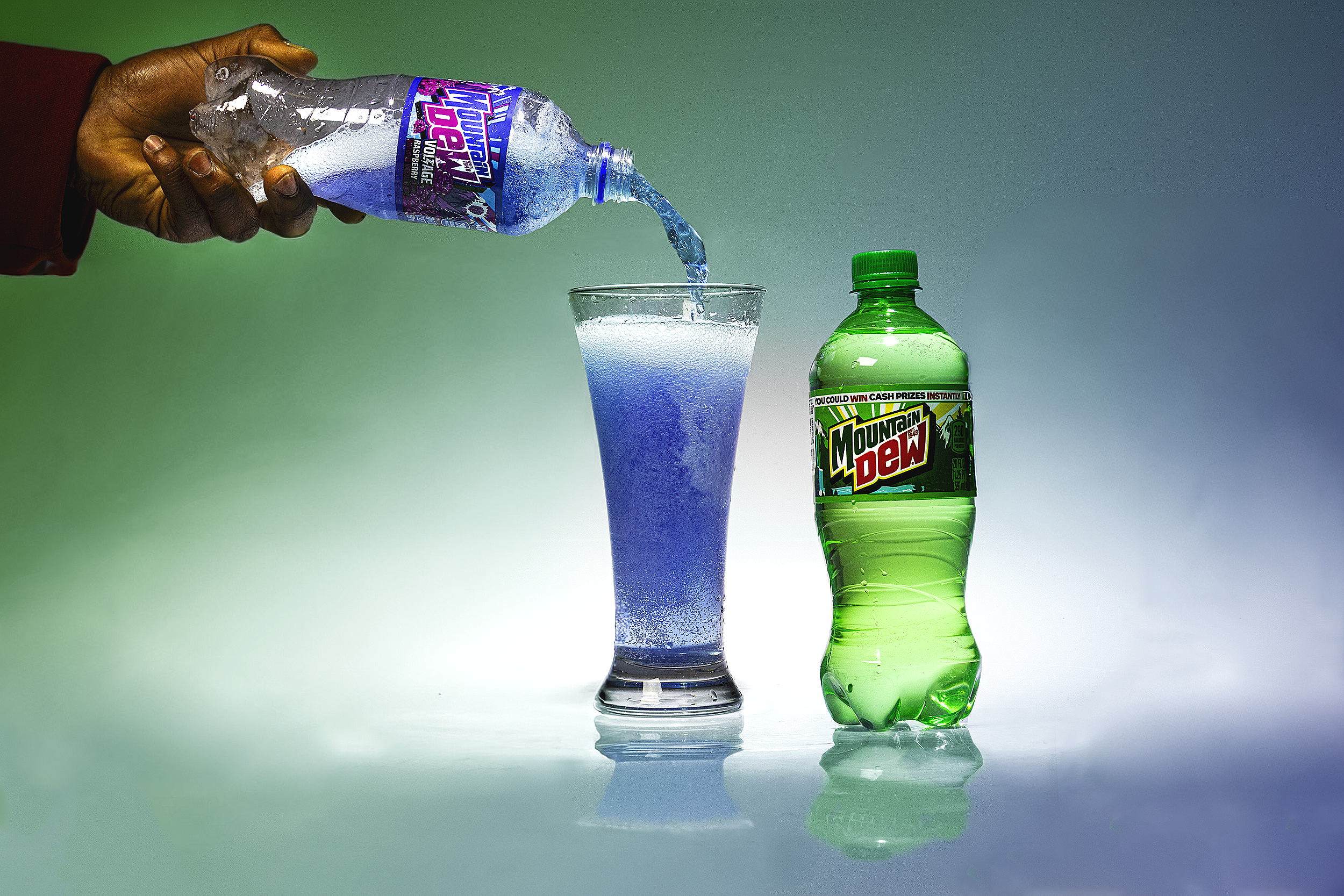 A person with dark skin and wearing a red sleeve is pouring Mountain Dew soda from a plastic bottle into a tall glass, with a second Mountain Dew bottle placed beside the glass. The background is a gradient green and gray.