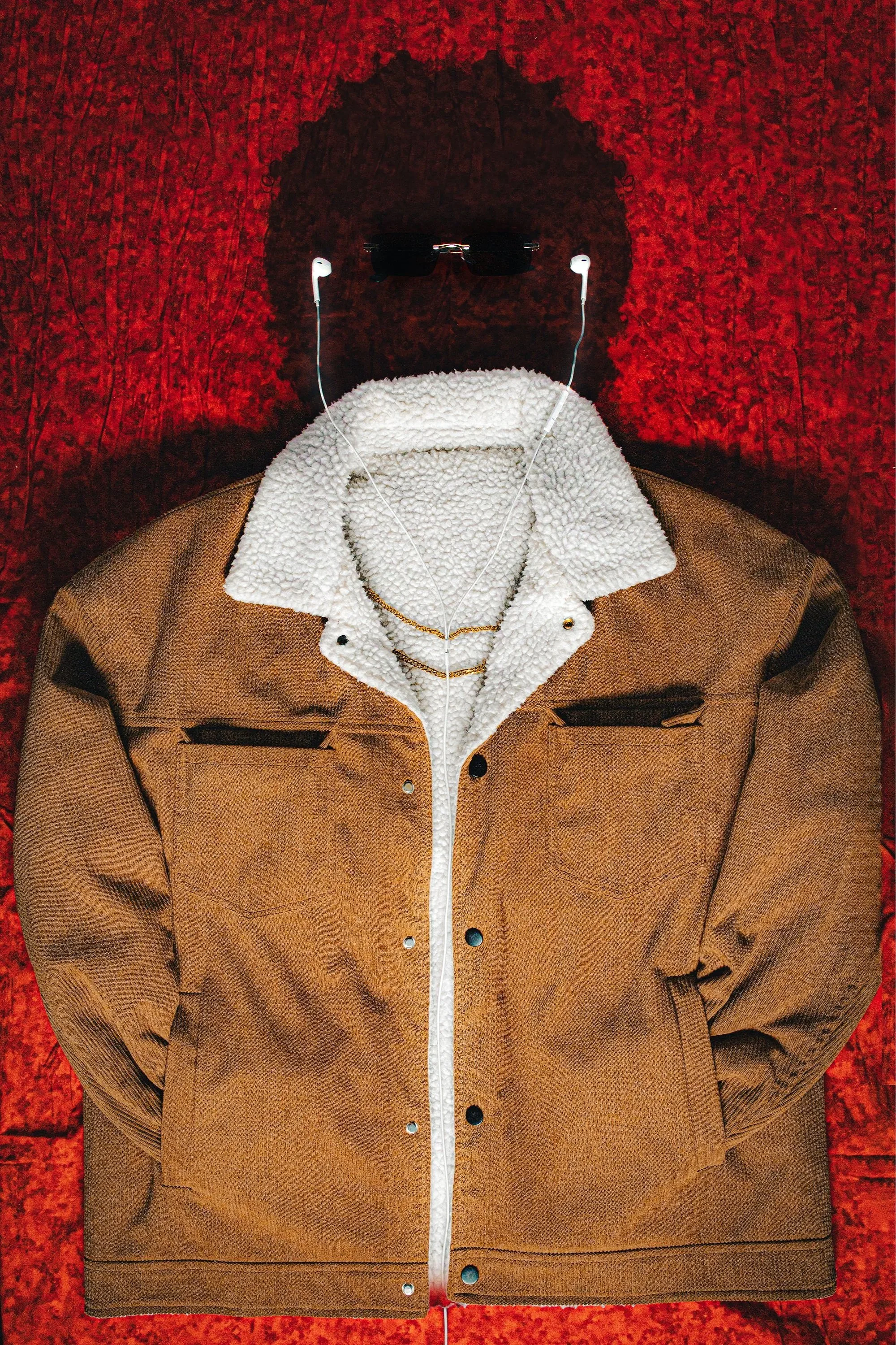 Person wearing sunglasses and white earphones, a white zip-up fleece, and a brown jacket, standing against a red textured background.