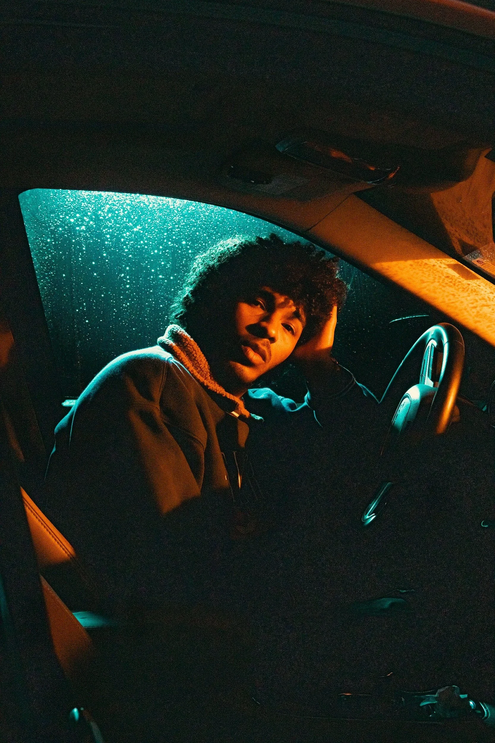A person with curly hair and a relaxed expression sitting inside a car at night, with rain visible on the windshield and illuminated by warm, orange interior lighting.
