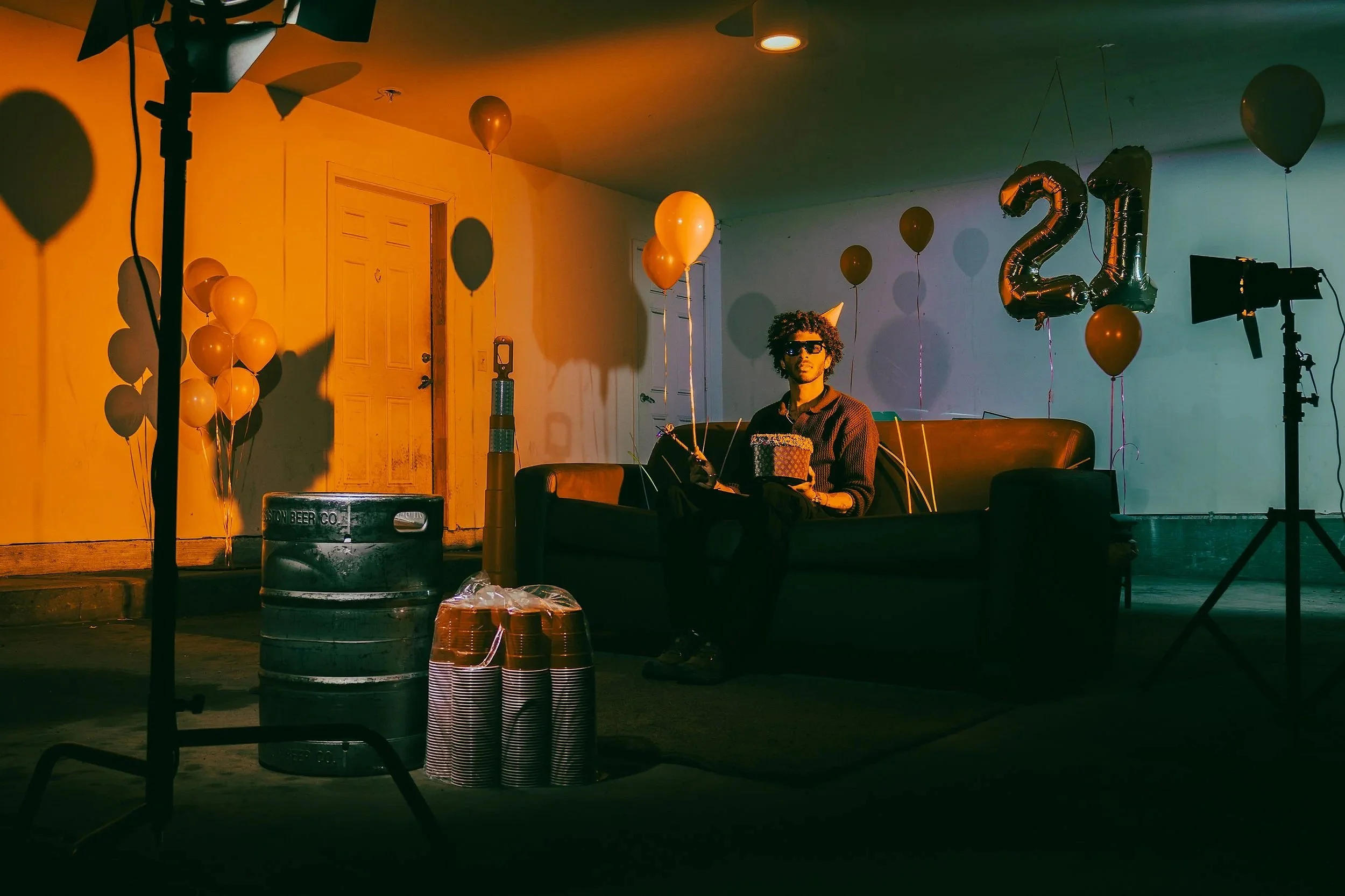 Young man with curly hair and sunglasses sitting on a couch, holding a Cupcake, surrounded by balloons, party hats, and large balloon numbers '2021', in a party setting with lighting and decorative props.