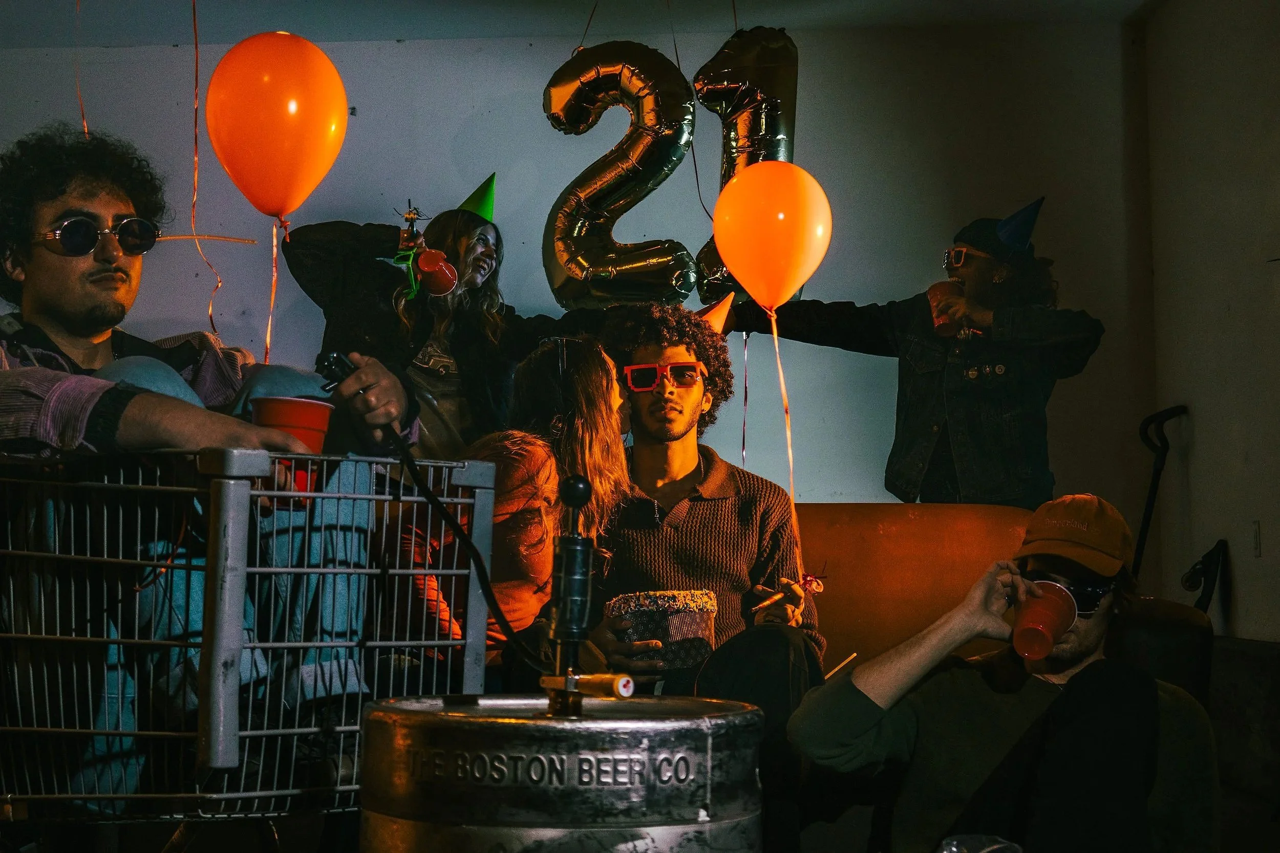 People celebrating a 21st birthday with balloons, party hats, and drinks in a dimly lit indoor setting.