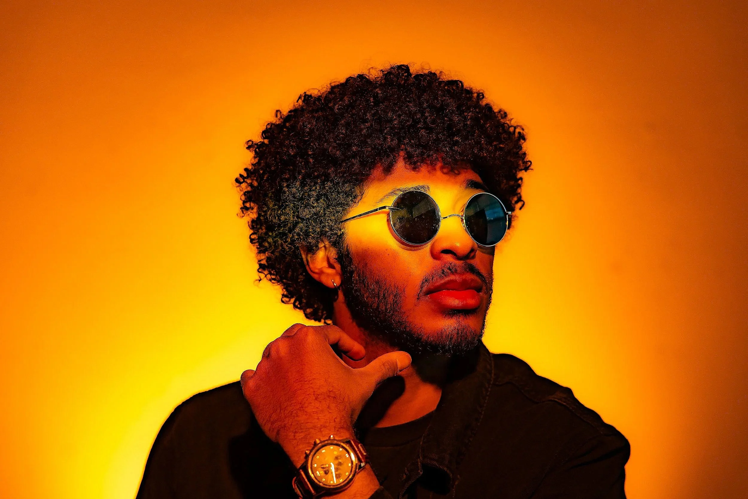 A young man with curly hair, sunglasses, and a beard is posing against an orange background. He is wearing a black shirt and a watch on his wrist, with his hand resting near his neck.
