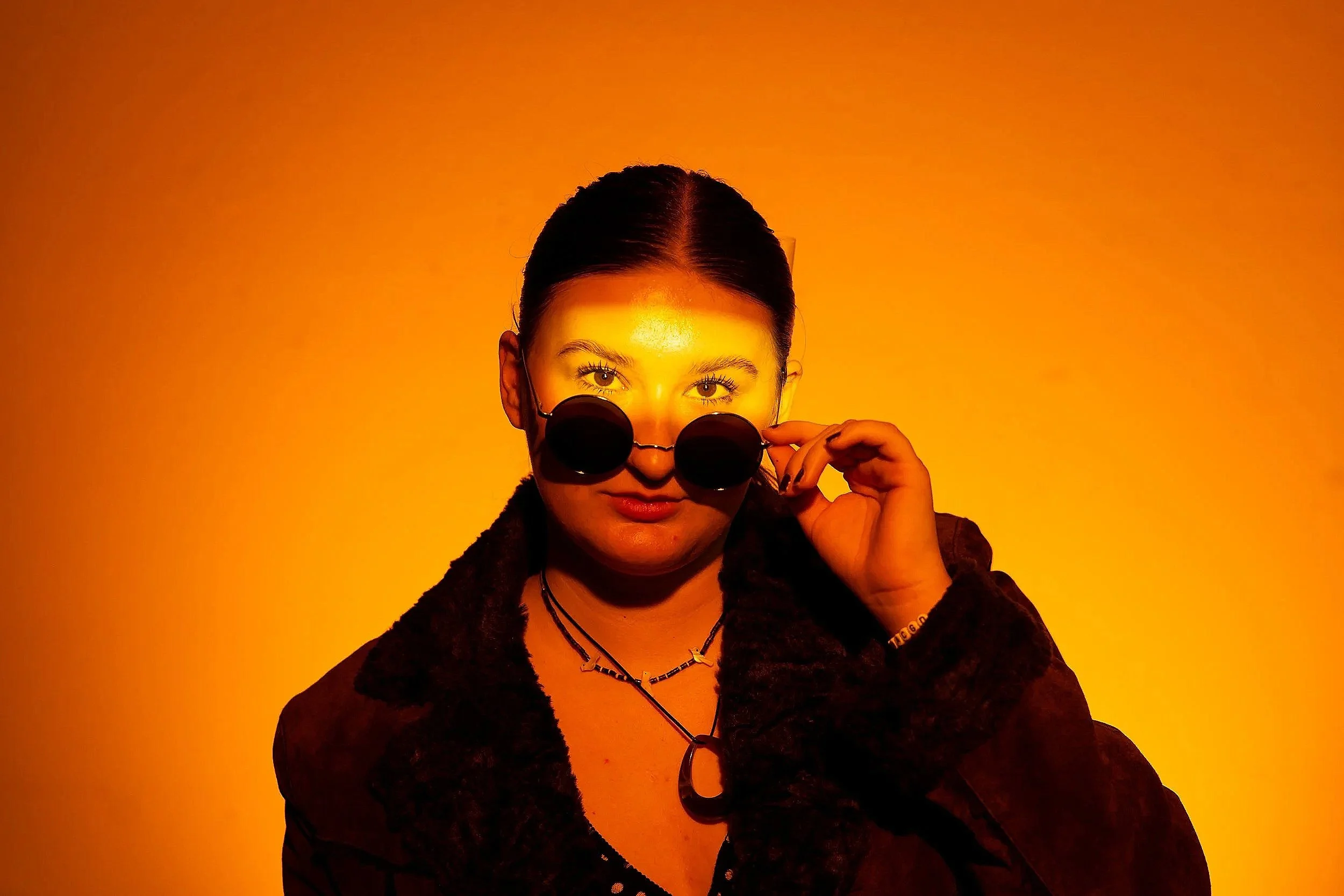 A woman with dark hair, wearing round dark sunglasses, a black top, and jewelry, is adjusting her sunglasses against a vibrant orange background.