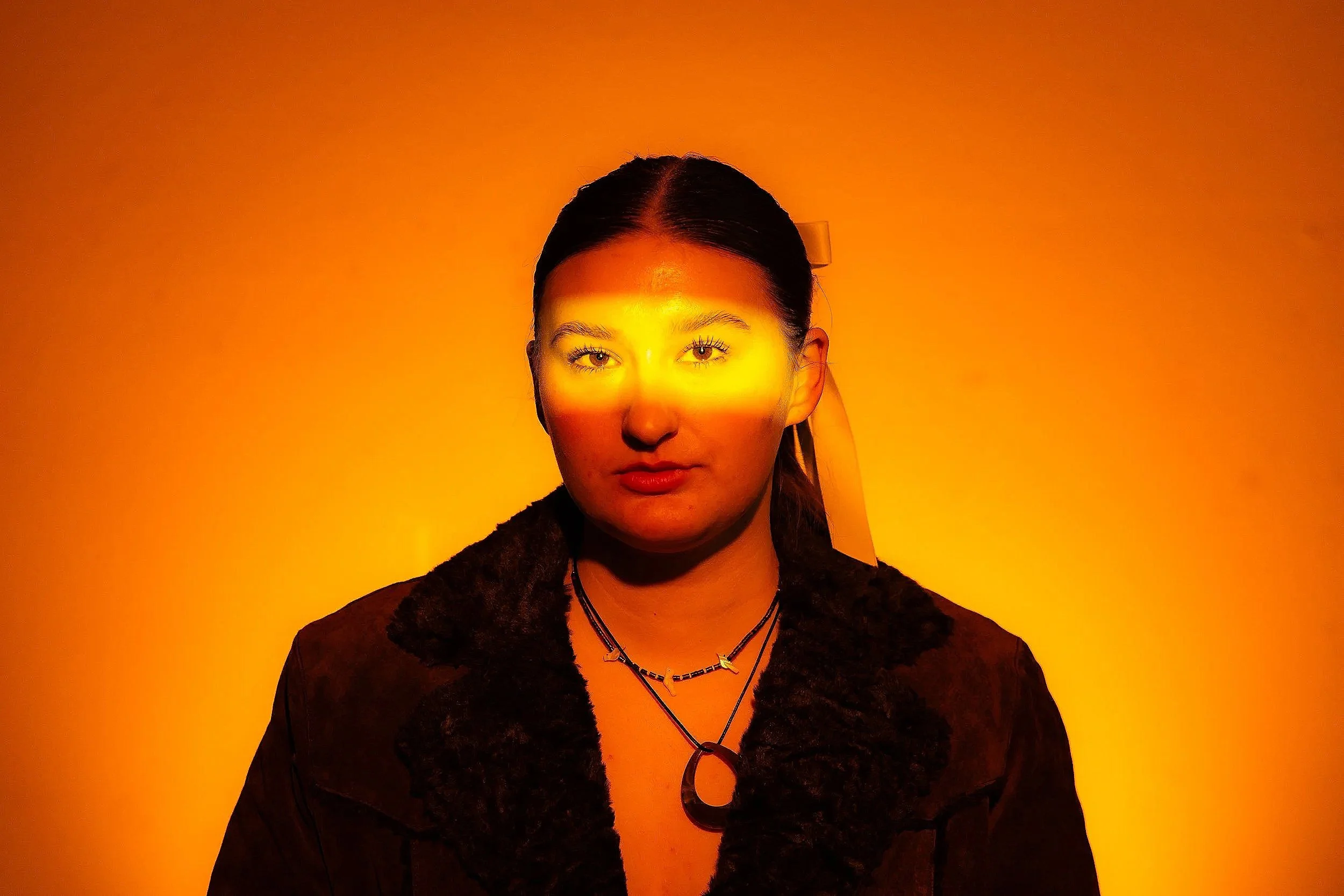 A woman with dark hair tied back, black and brown clothing, and multiple necklaces, standing against a bright yellow-orange background with a curved lighting pattern across her face.
