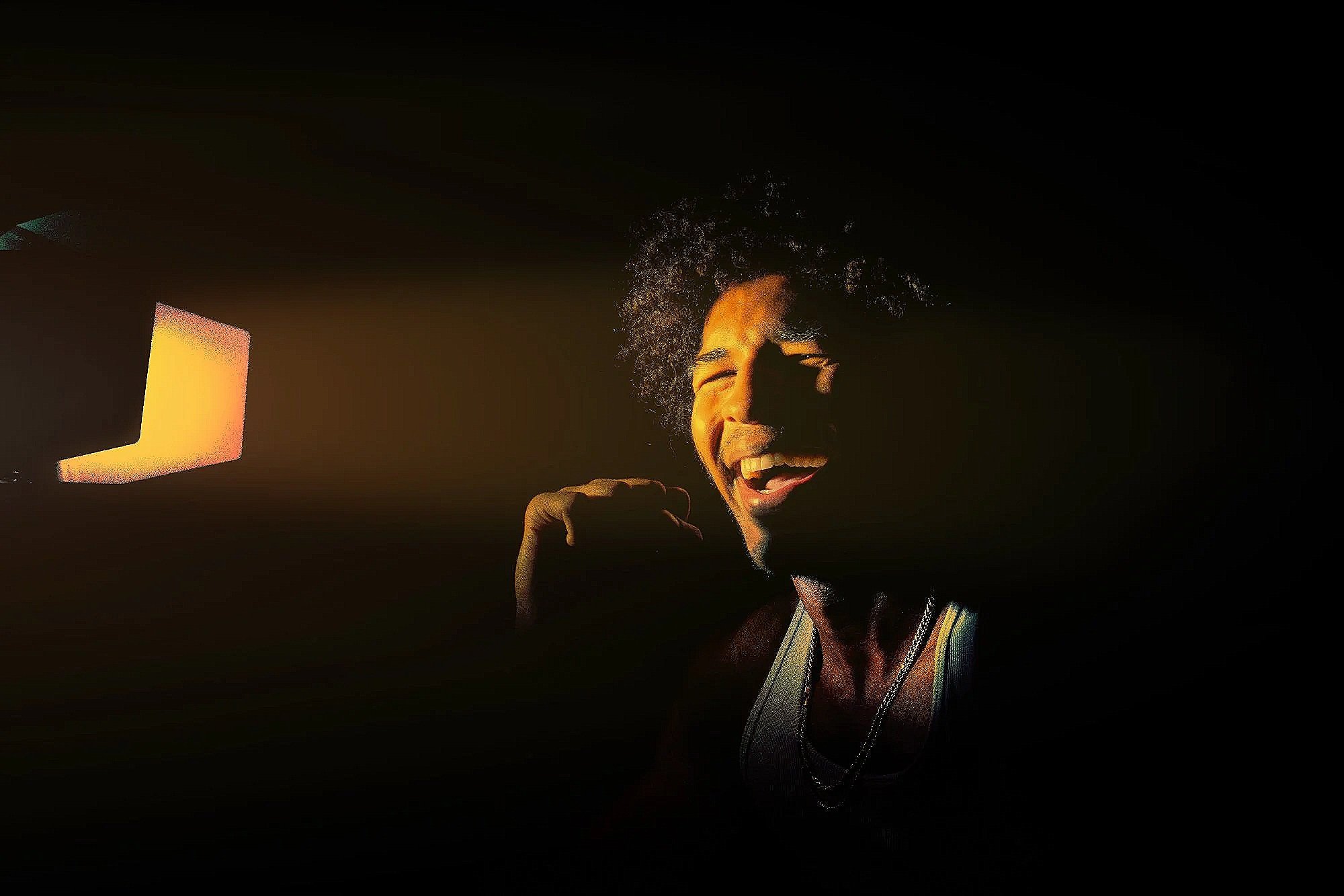 A person with curly hair smiling and singing into a microphone, illuminated by a screen on the left, in a dark environment.