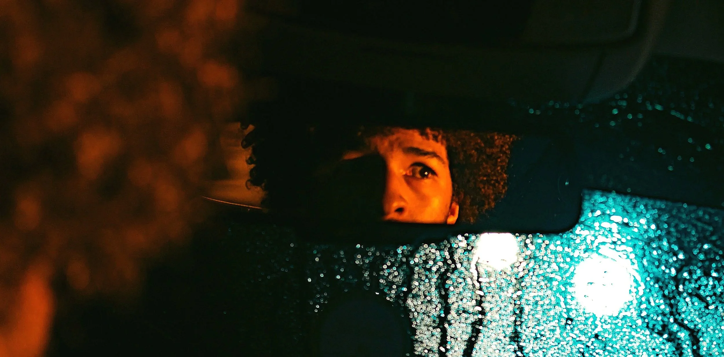 A man with curly hair looking into a wet rearview mirror during rain at night, illuminated by streetlights.