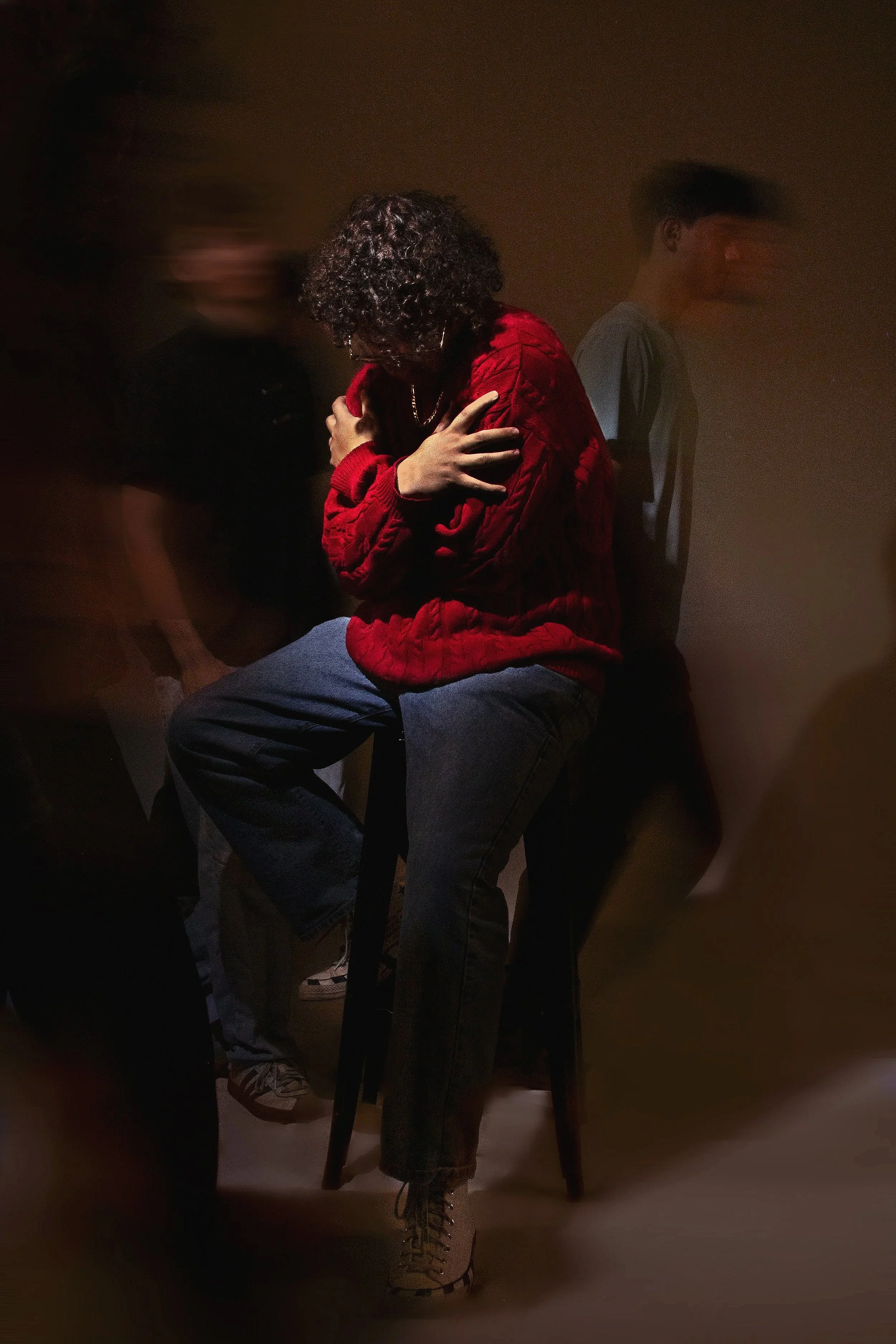 A woman with curly hair wearing a red jacket and jeans, sitting on a black chair with her arms crossed over her chest, in a dimly lit room with two other people nearby.