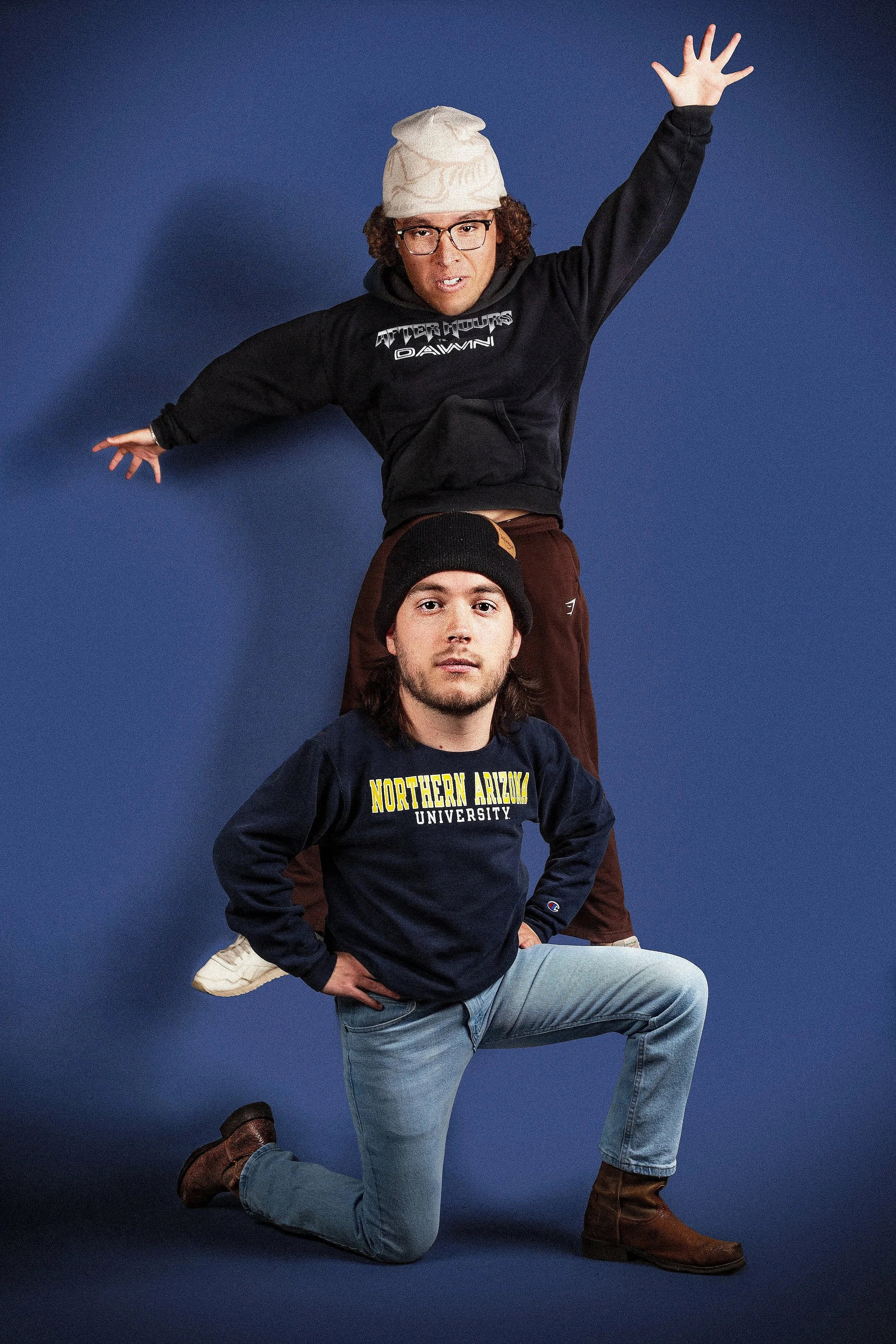 Two men posing against a dark blue background. The man at the top is standing on the shoulders of the man below him. The man at the top is wearing glasses, a white beanie, a black hoodie with logos, and brown pants. The man below is kneeling with one