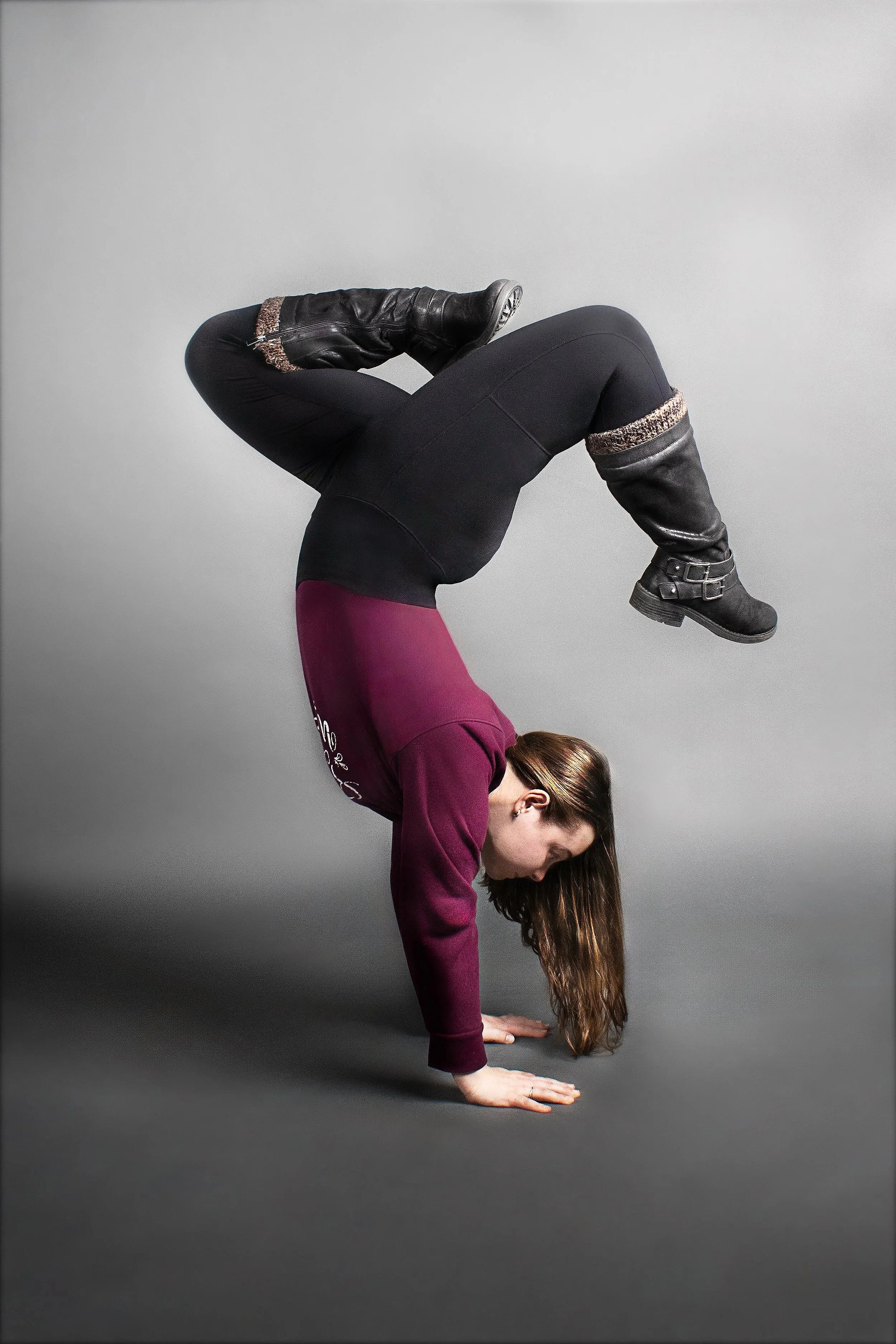 A woman performing a yoga pose in a handstand position, supporting herself with her hands on the ground, her legs bent at the knees, and her head lowered toward the ground against a plain gray background.