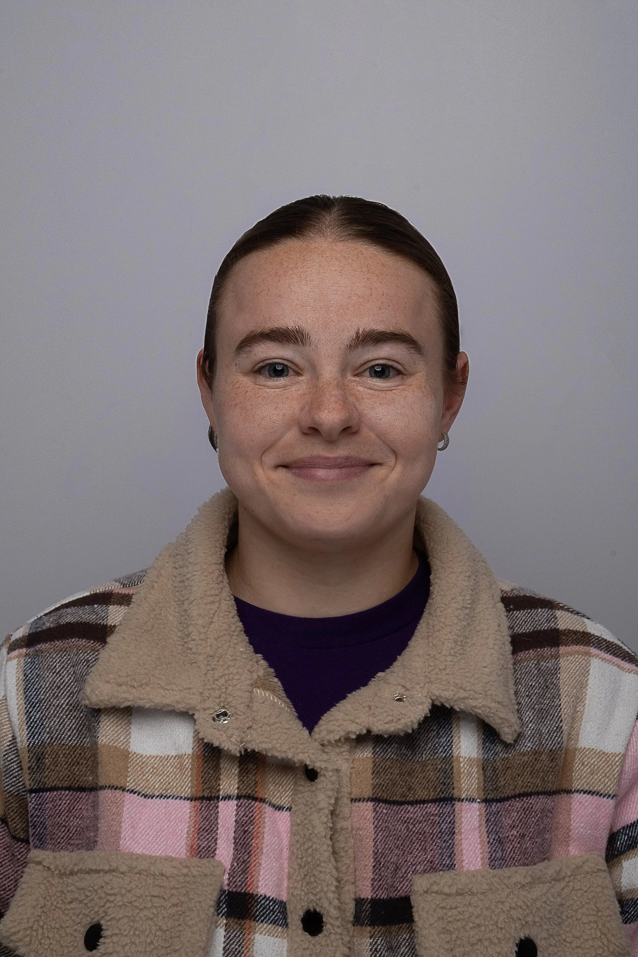 A person with fair skin and short, dark hair smiling at the camera. They are wearing a plaid jacket with beige, pink, brown, and black colors, a dark shirt underneath, and small hoop earrings. The background is a plain light gray wall.