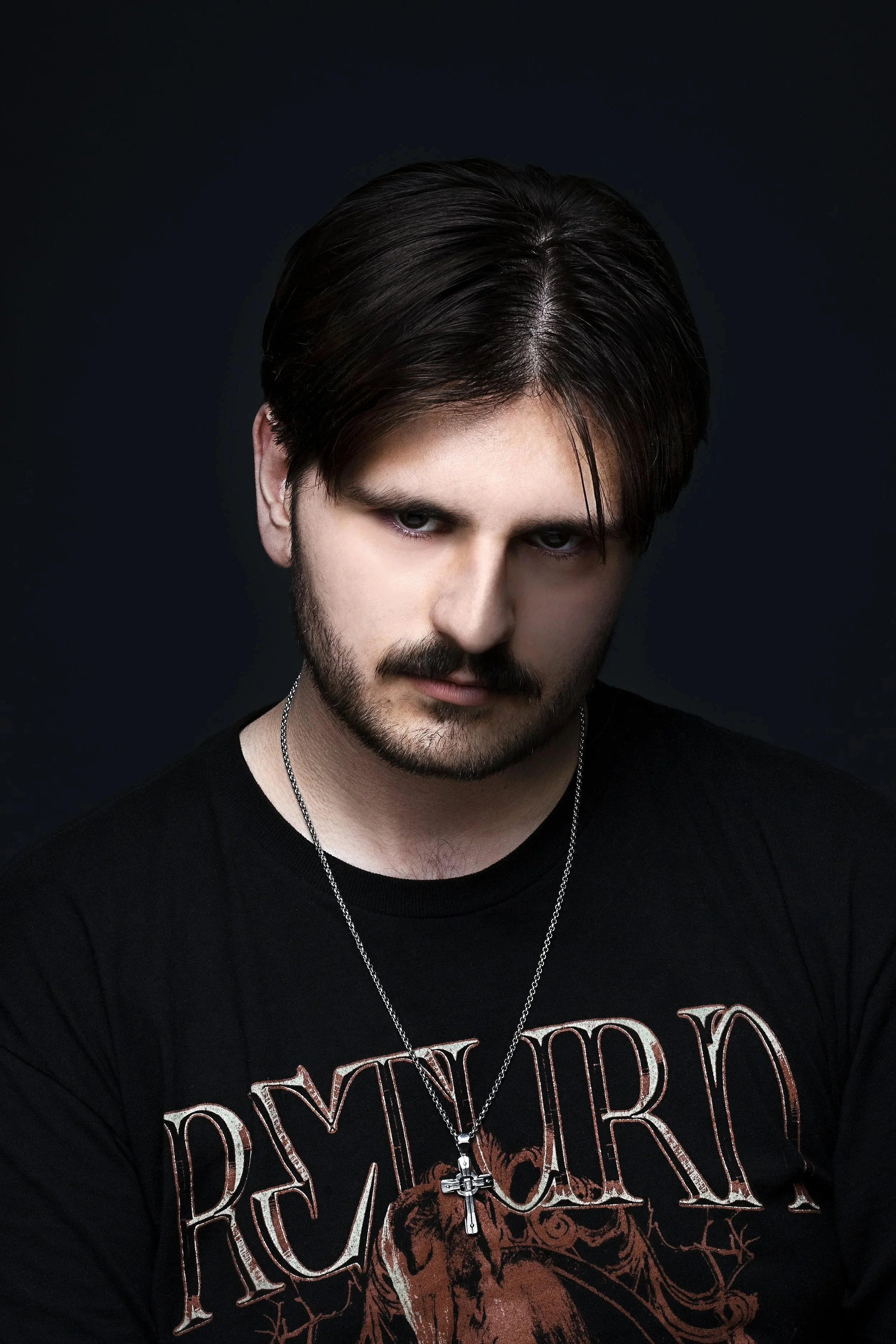 A young man with dark hair and facial hair wearing a black graphic T-shirt and a chain necklace with a cross pendant, looking directly at the camera against a dark background.
