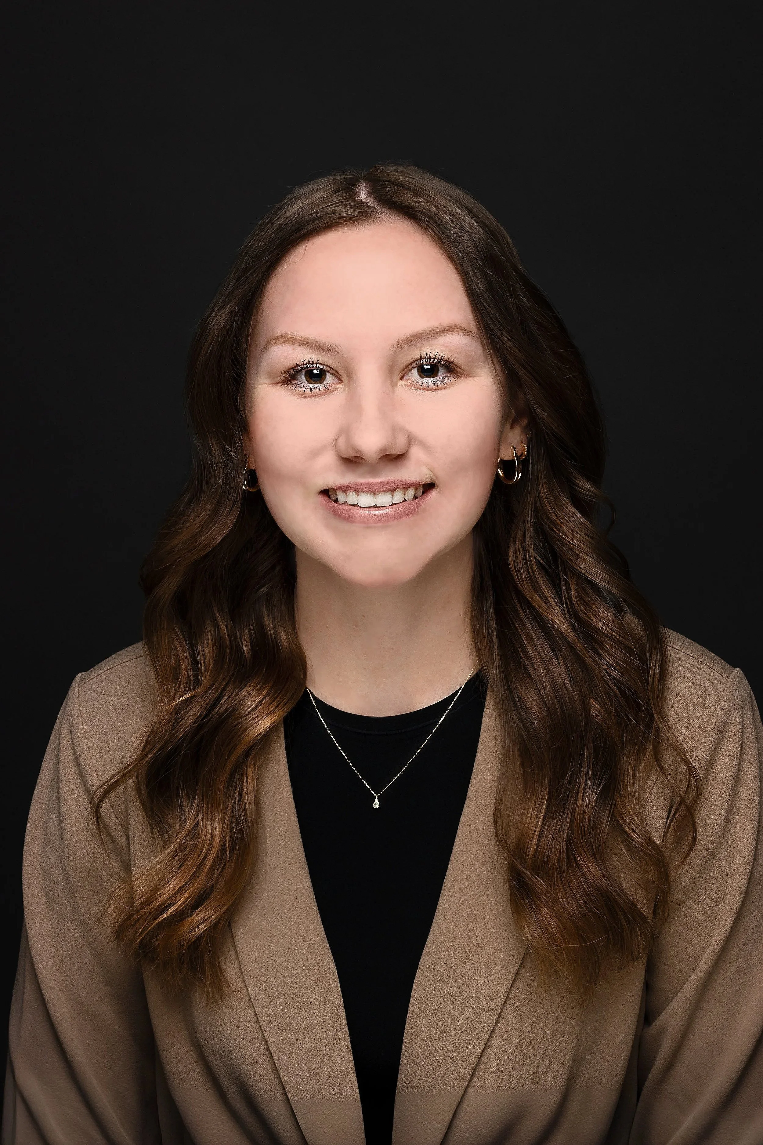 Portrait of a young woman with long wavy brown hair, wearing a beige blazer, black top, earrings, and a necklace, smiling against a black background.