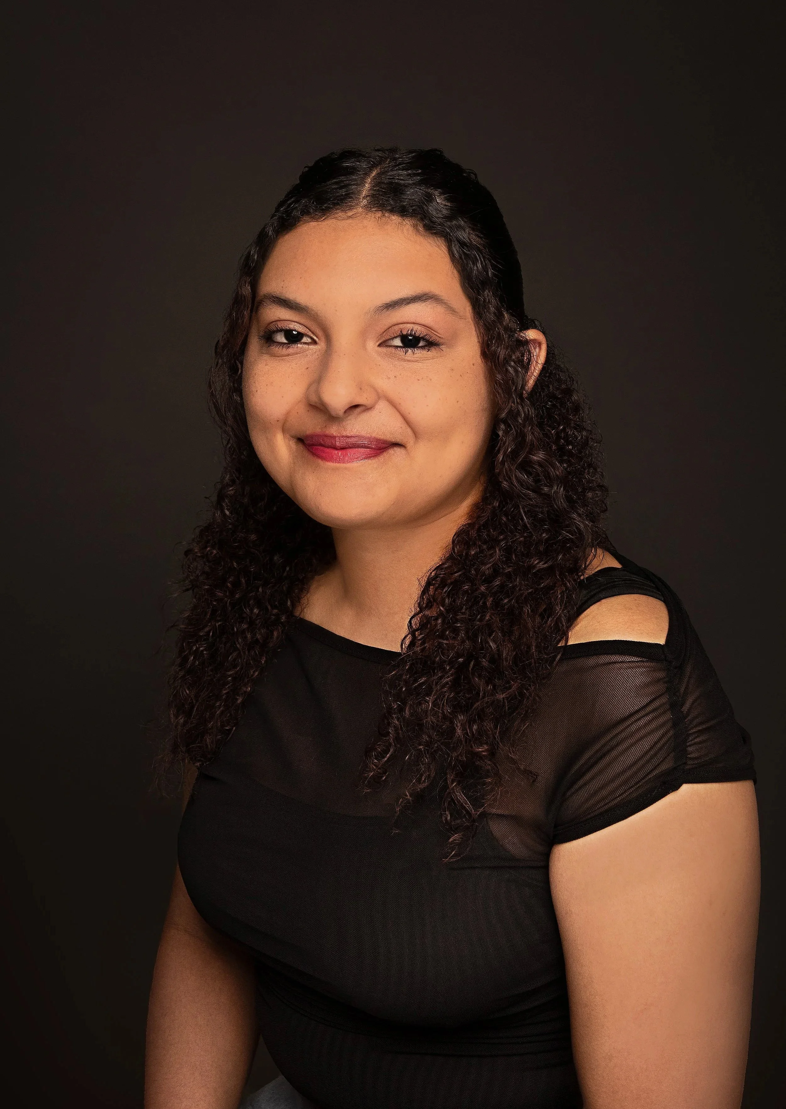 A young woman with curly brown hair, wearing a black top with sheer sleeves, smiling softly against a dark background.