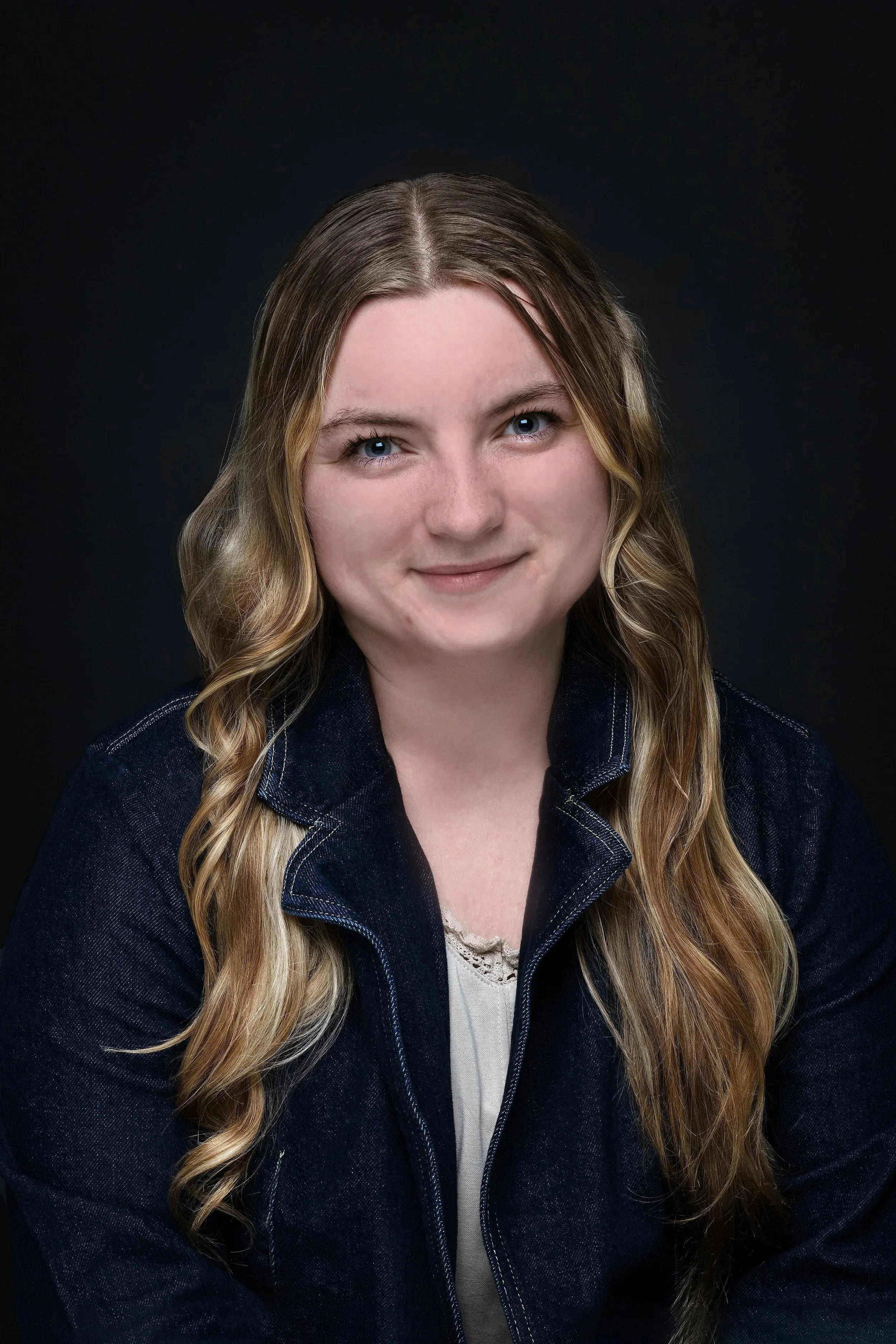 A young woman with long, wavy blonde hair and blue eyes smiling against a dark background, wearing a denim jacket over a light-colored top.