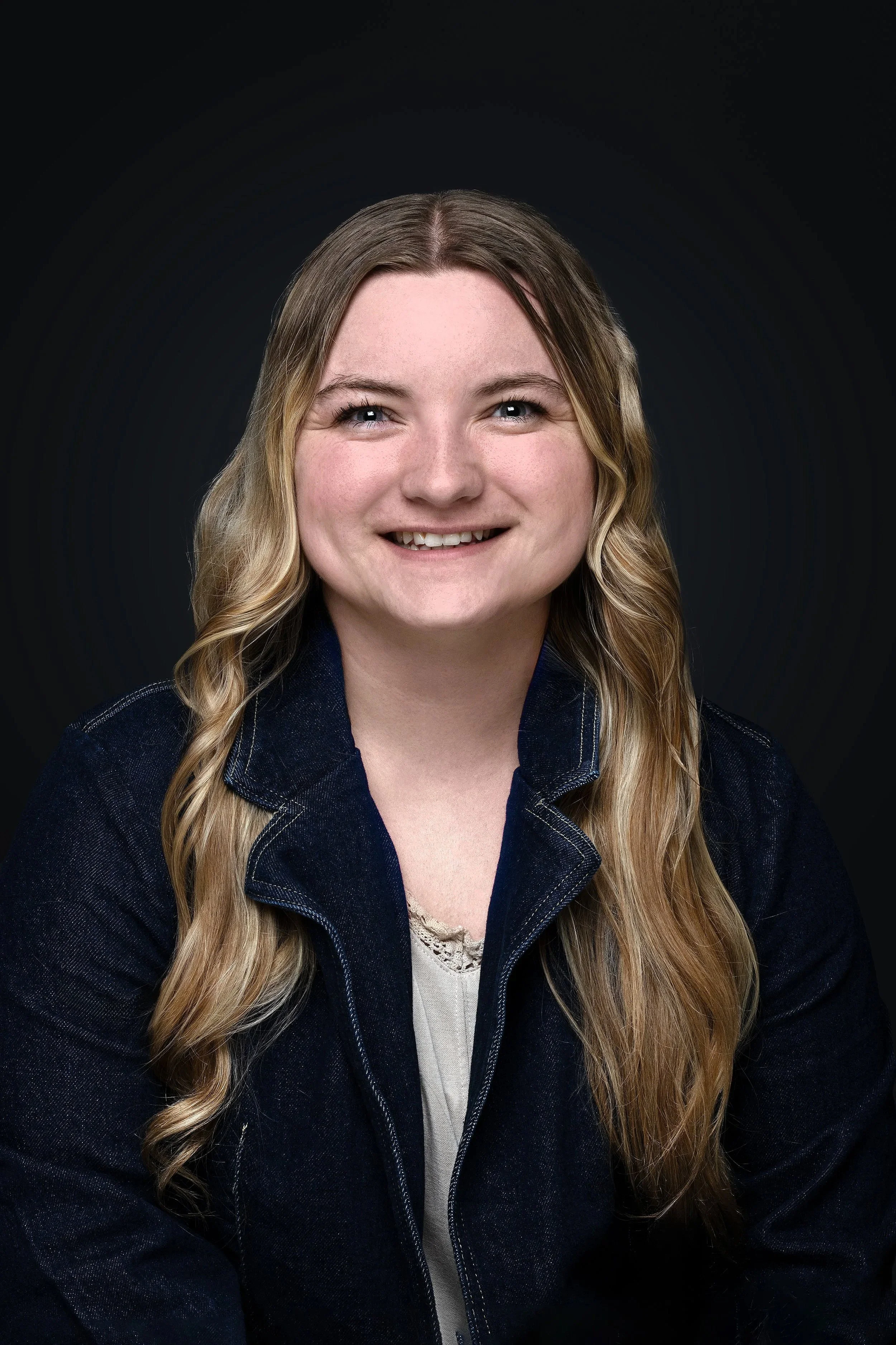 Close-up portrait of a woman with long wavy blond hair, smiling, wearing a dark denim jacket and a white lace top, set against a dark background.