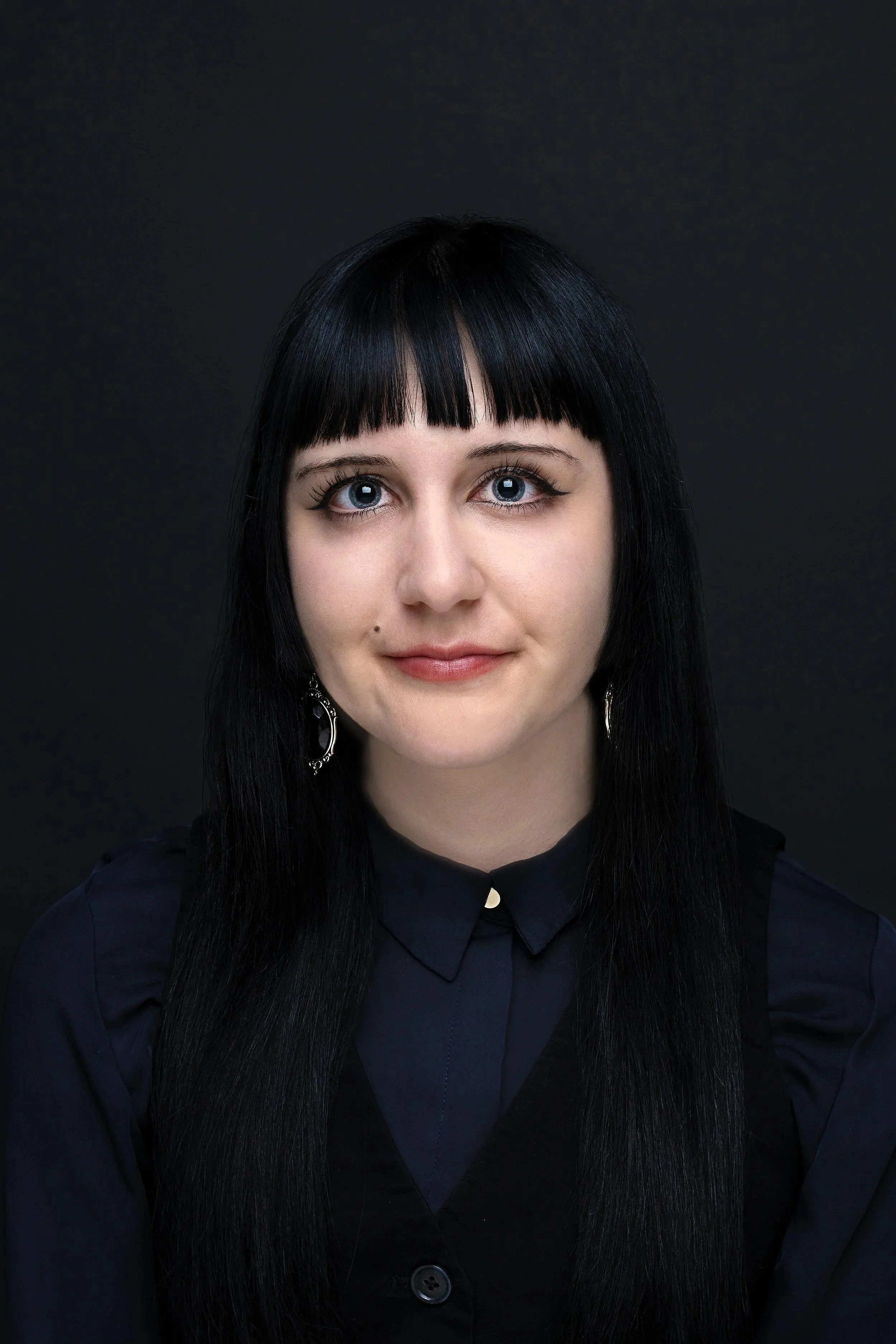 Portrait of a woman with long black hair and blunt bangs, wearing a navy blue shirt with a collar, silver earrings, and set against a dark background.