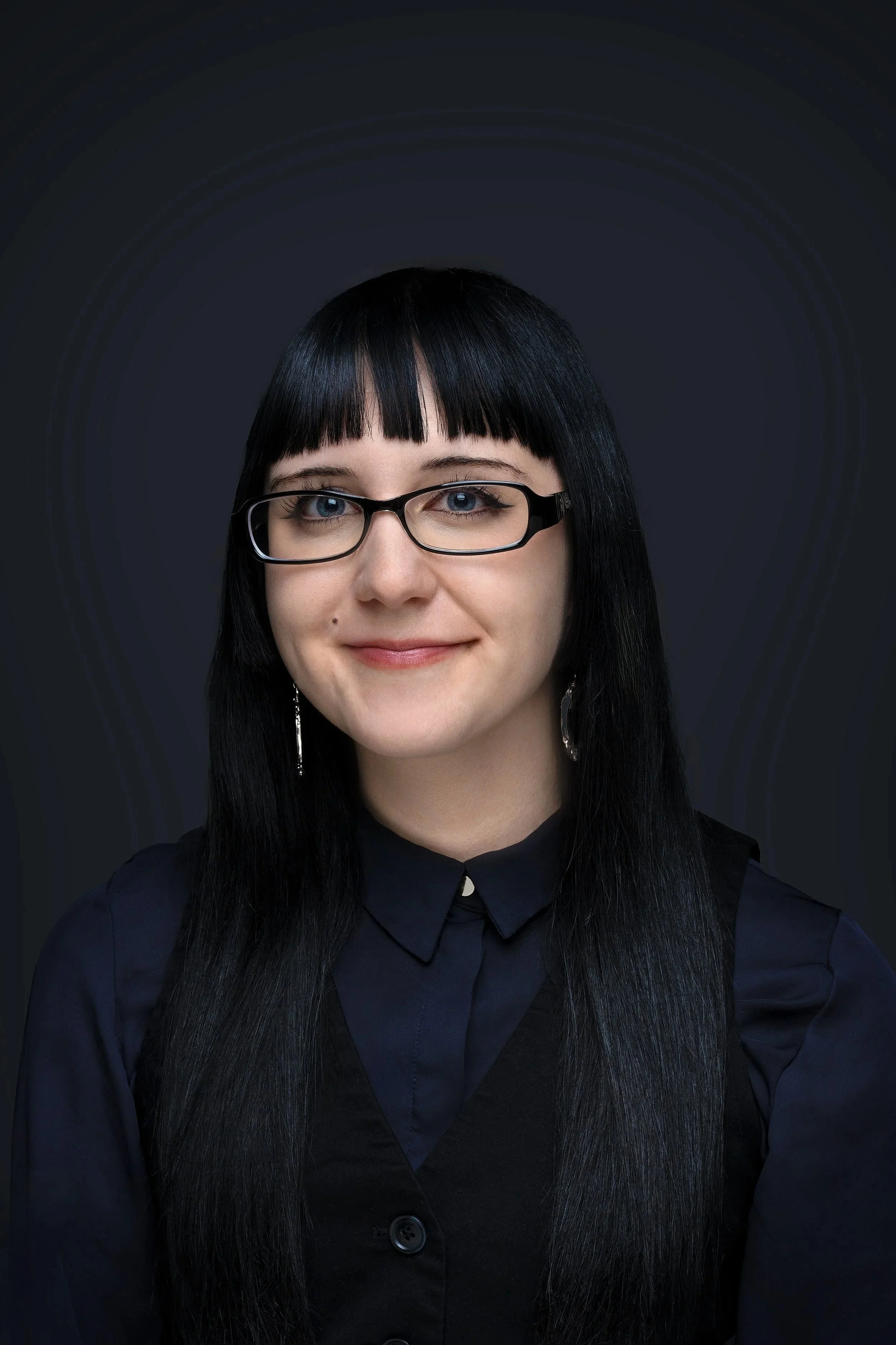 Portrait of a woman with long black hair, wearing black glasses and a dark button-up shirt, smiling against a dark background.