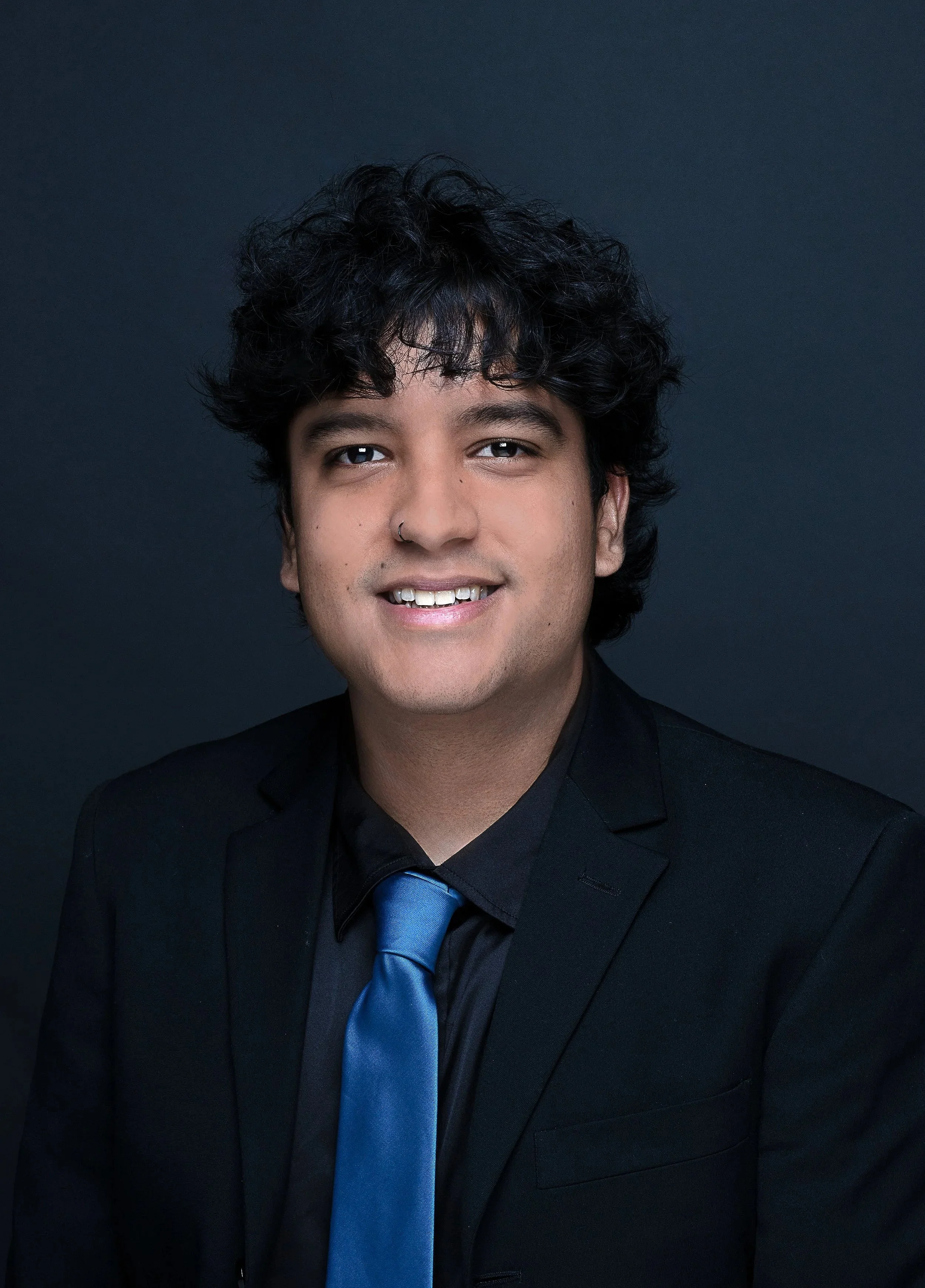 Portrait of a young man with black curly hair, wearing a black suit and blue tie, smiling against a dark background.