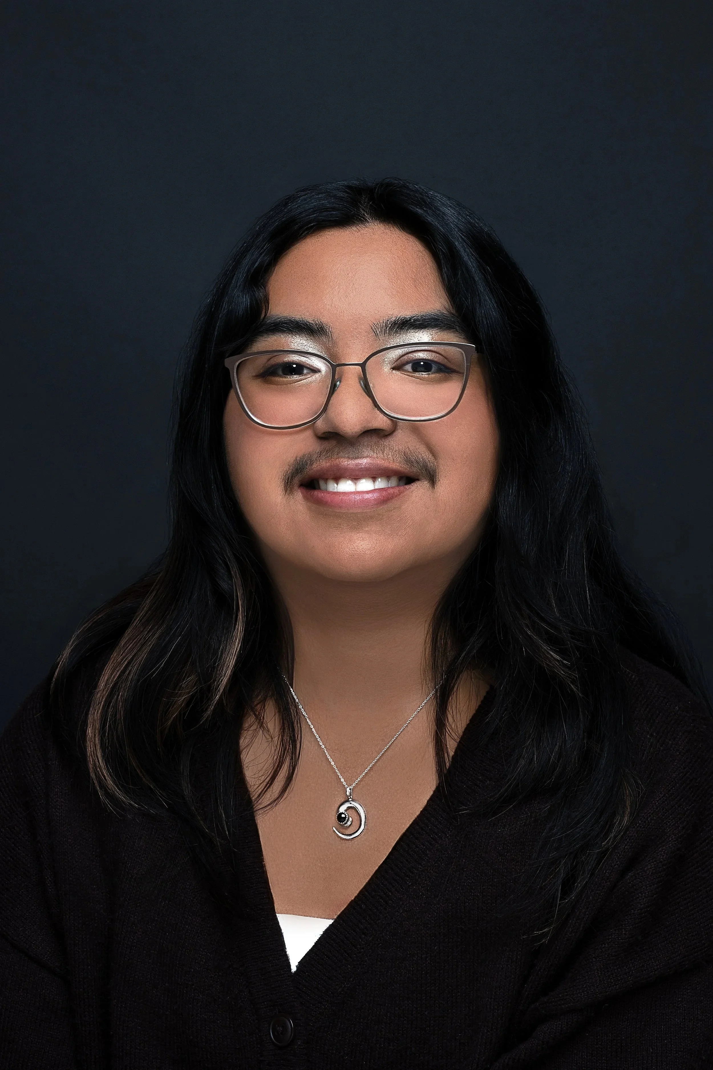 Close-up portrait of a smiling woman with long dark hair, glasses, and wearing a black cardigan and silver jewelry, against a dark background.