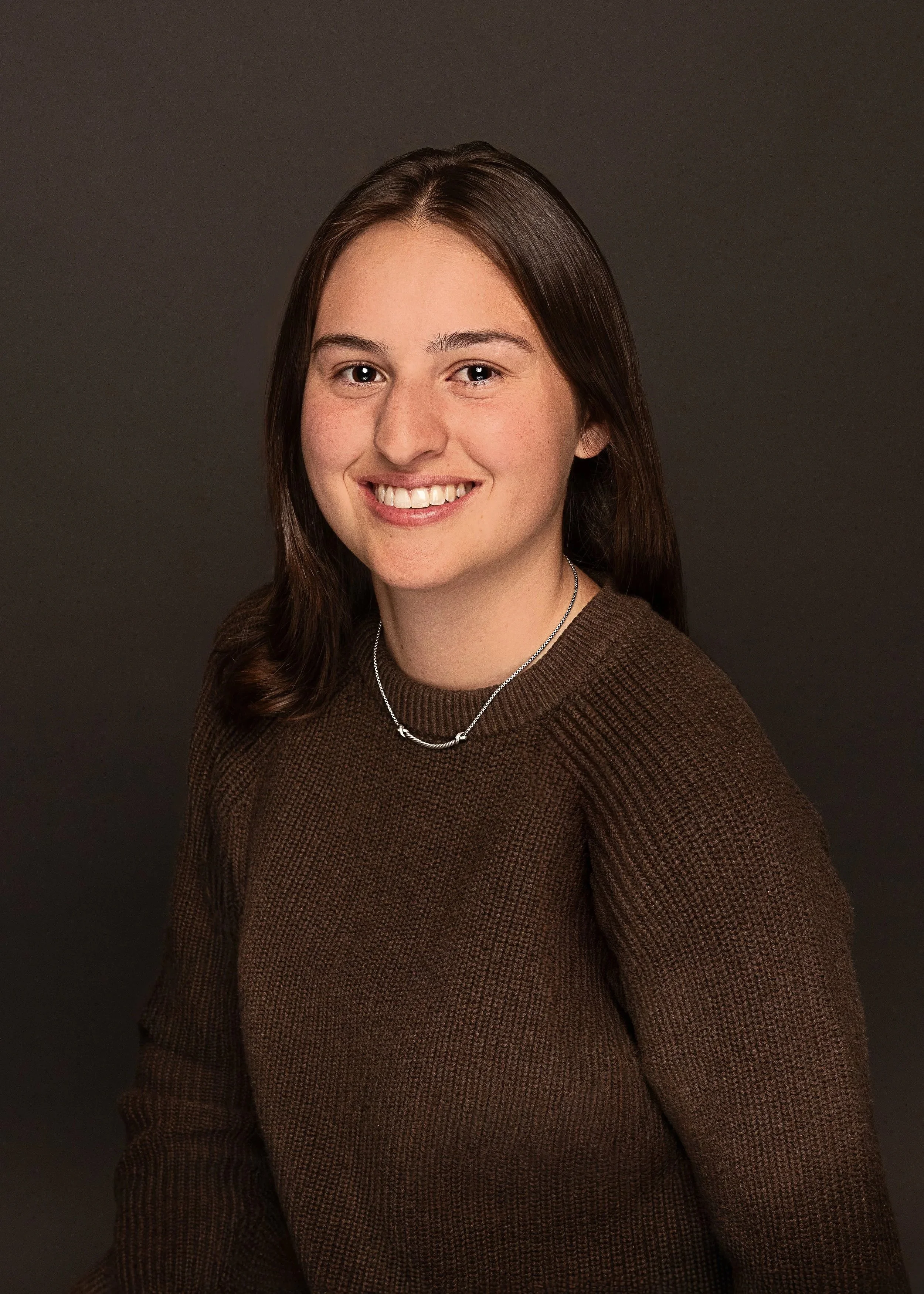 A young woman with long brown hair, wearing a brown sweater and a silver necklace, smiling against a dark background.