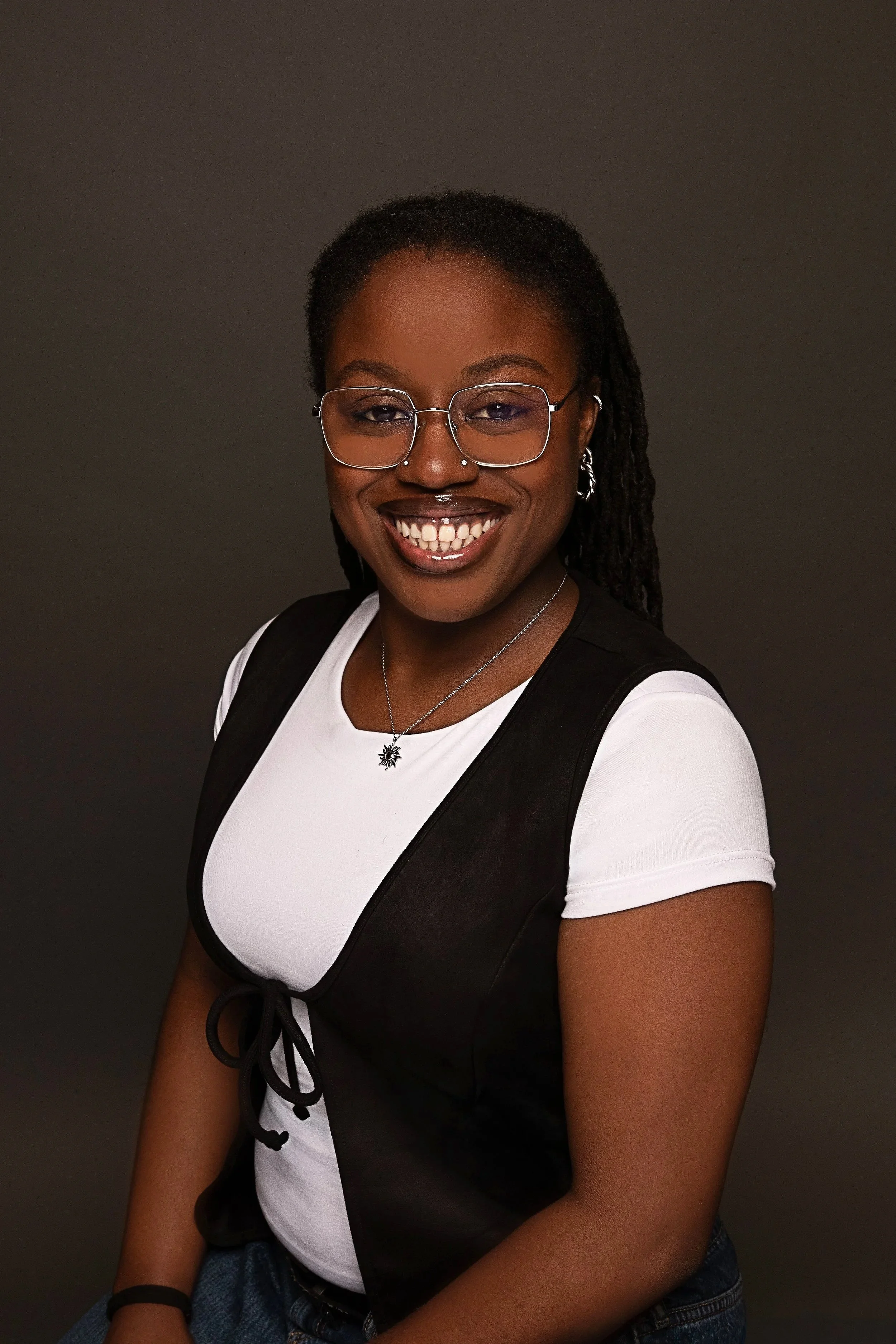 A woman with dark skin, glasses, and dreadlocked hair smiling at the camera. She is wearing a black and white shirt, a necklace, earrings, and carrying a backpack against a dark background.