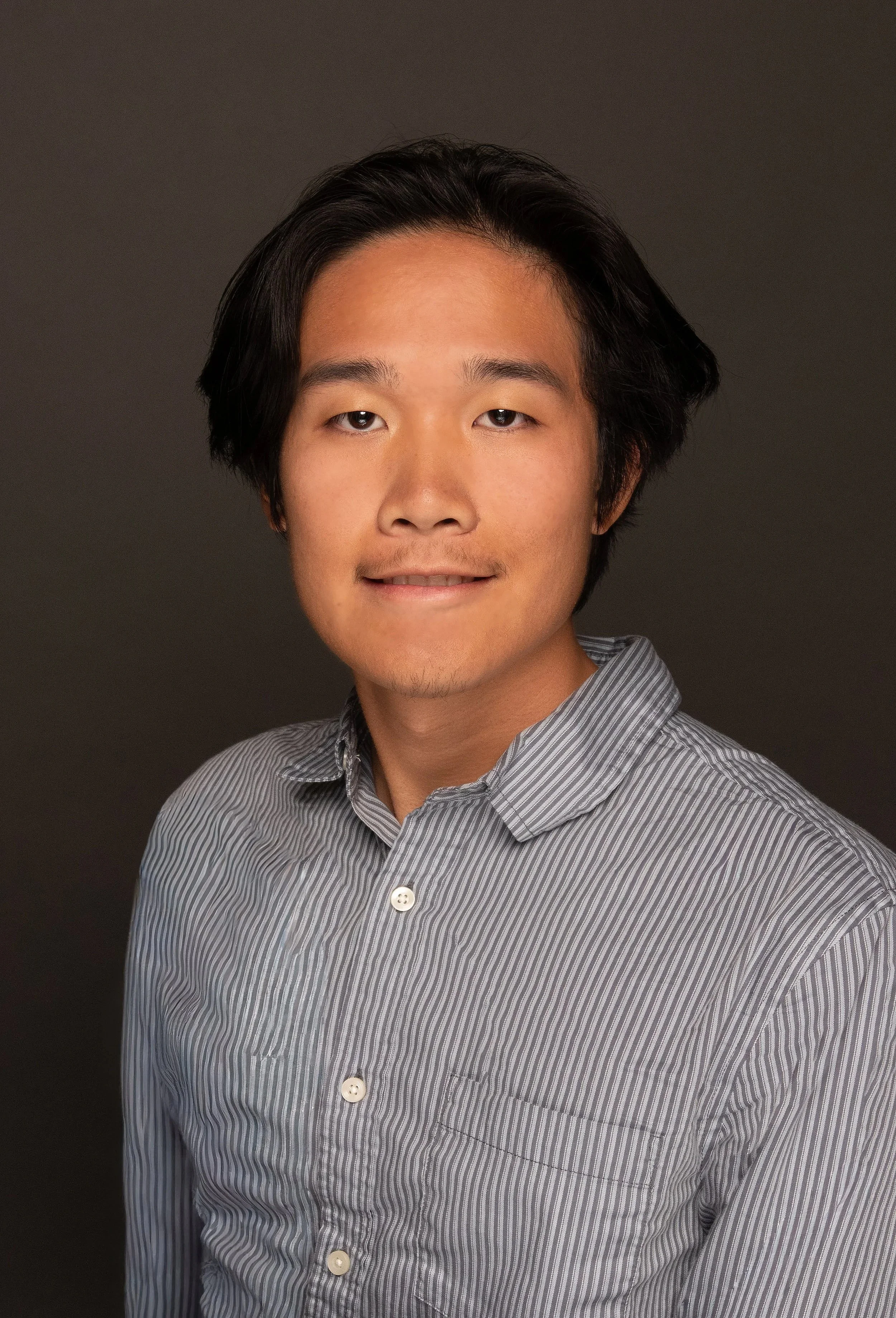 A young man with black hair and light brown skin wearing a gray striped shirt against a dark background.