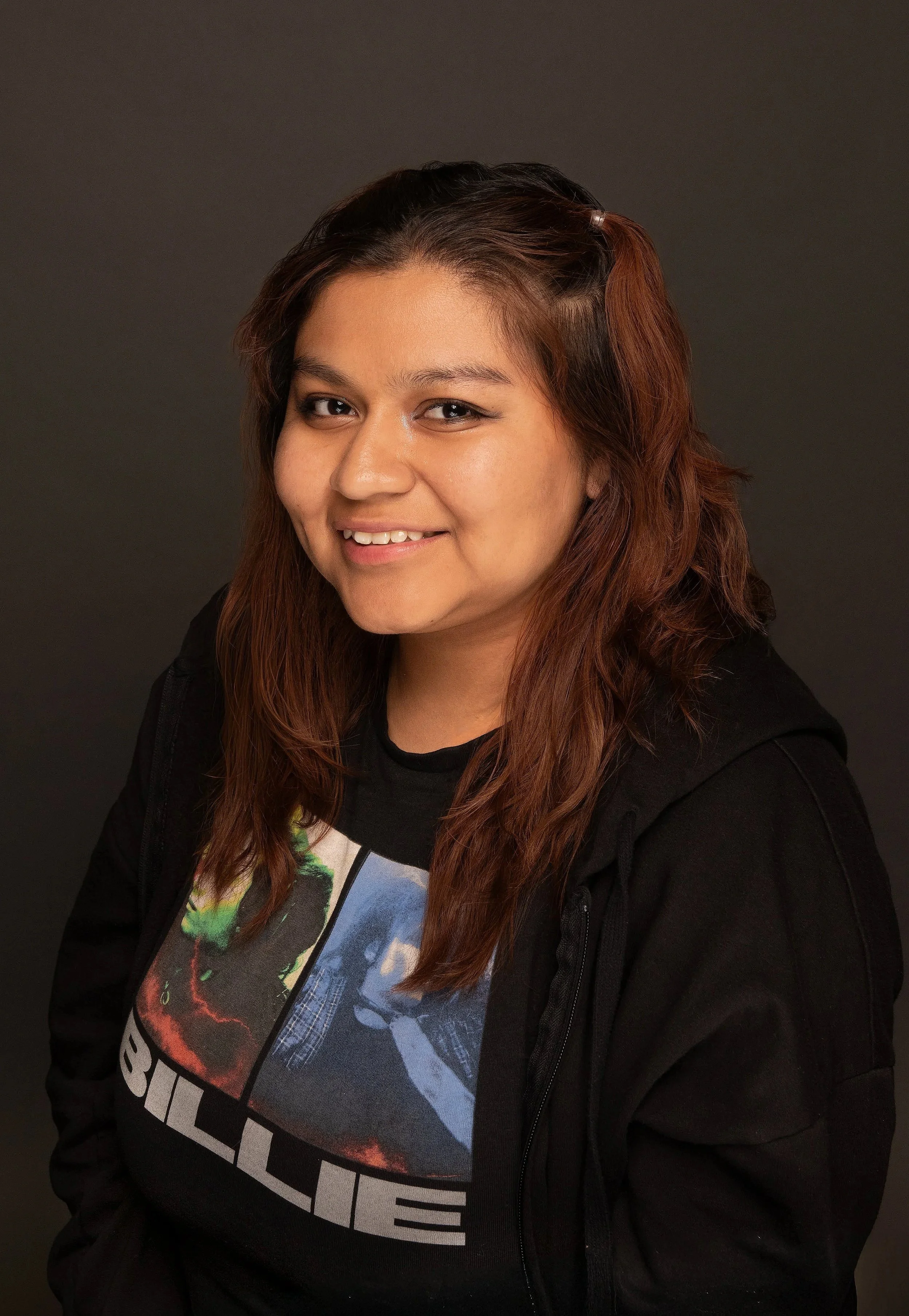 Portrait of a woman with long, wavy brown hair smiling, wearing a black jacket and a graphic T-shirt, against a plain dark background.