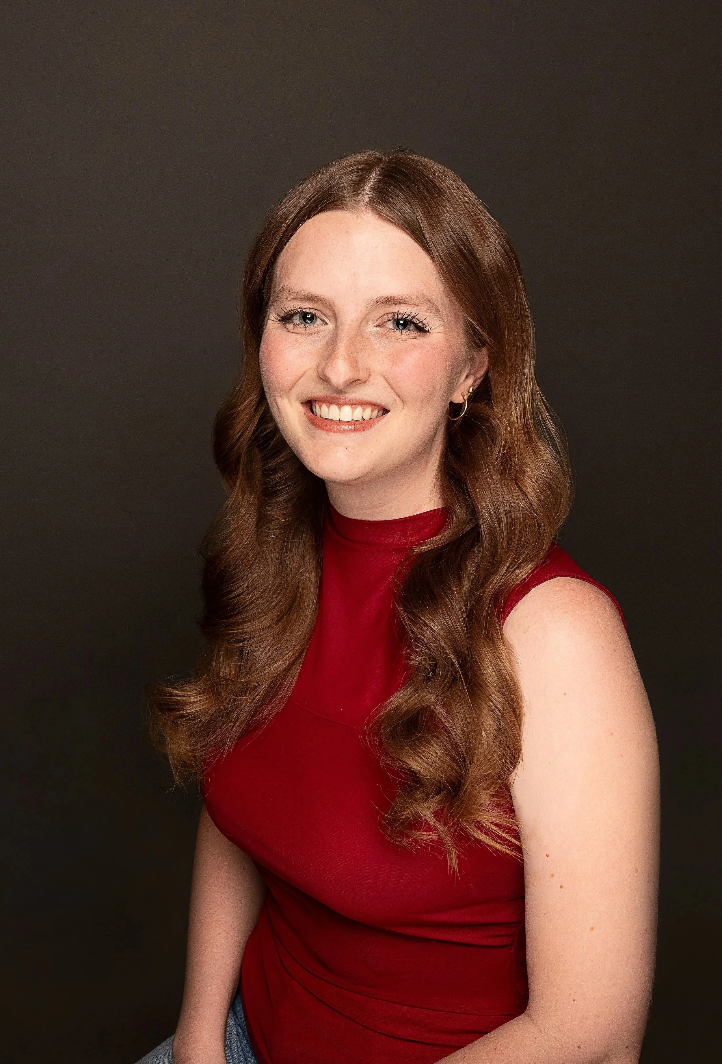 Portrait of a young woman with long, wavy auburn hair, wearing a red sleeveless top, smiling against a dark background.