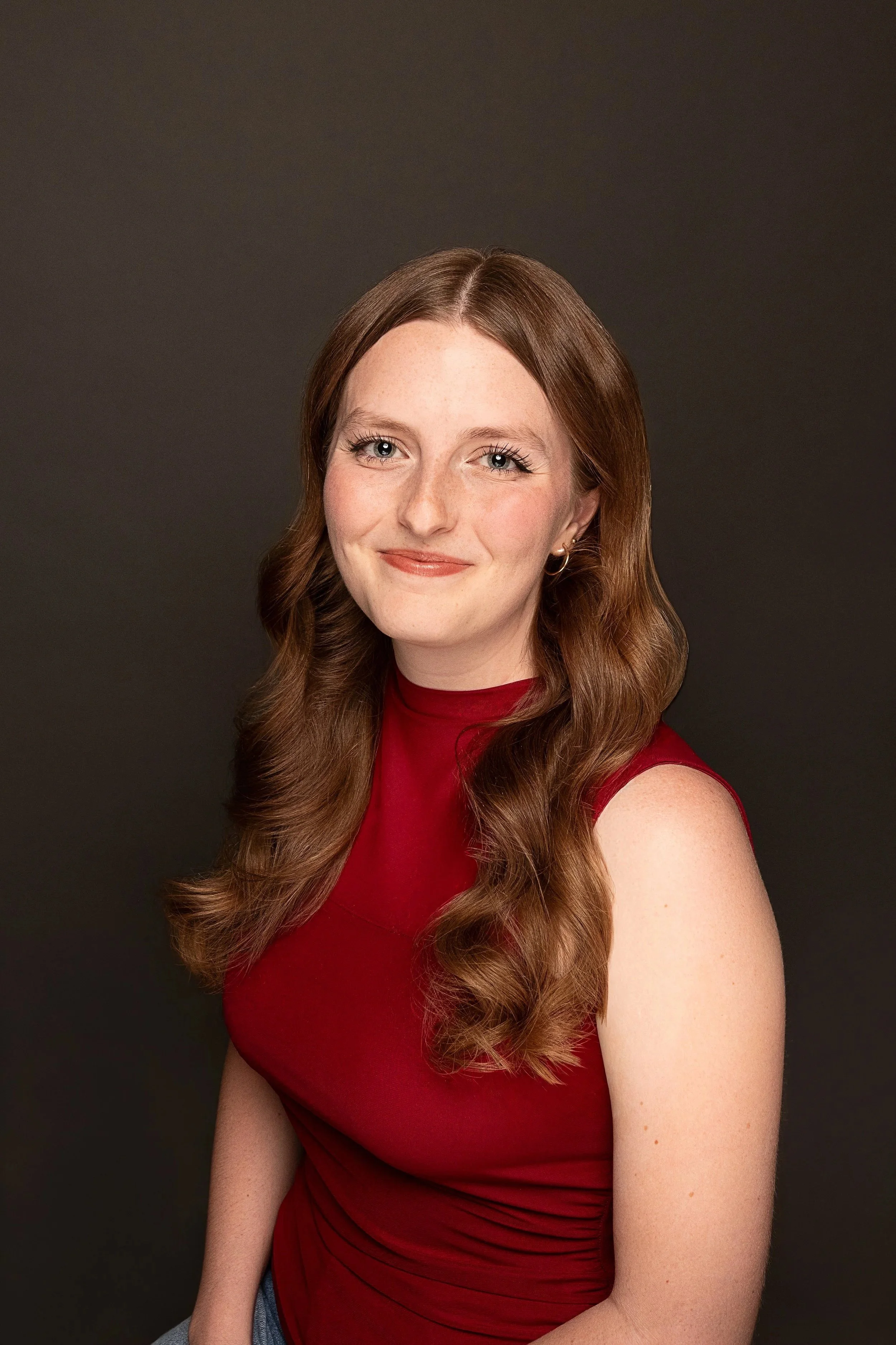 Portrait of a young woman with long wavy red hair, wearing a sleeveless red top, and silver hoop earrings, against a dark background.