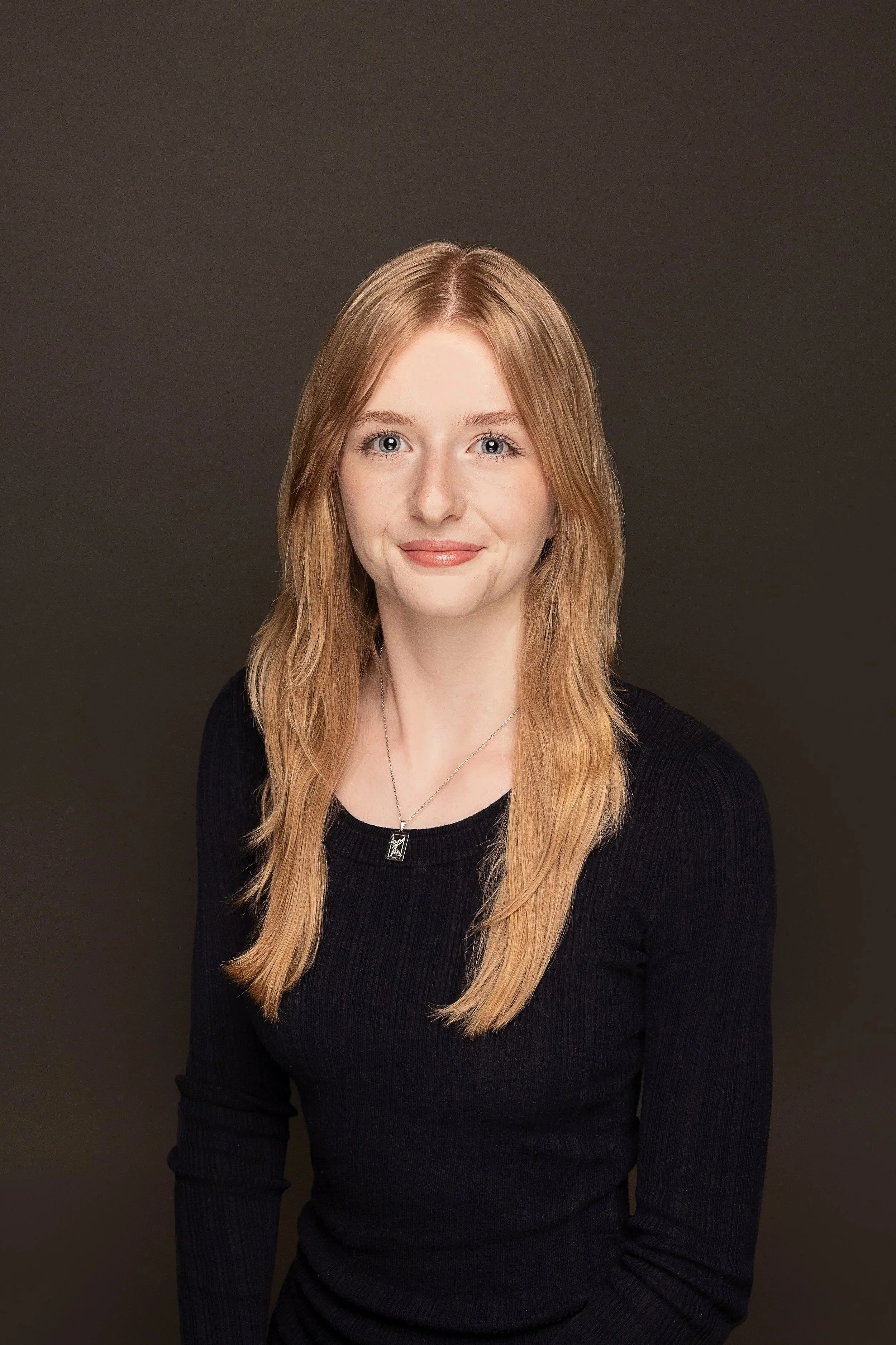 A young woman with long, reddish hair, blue eyes, and fair skin, wearing a black long-sleeve top and a silver necklace, smiling slightly against a dark background.