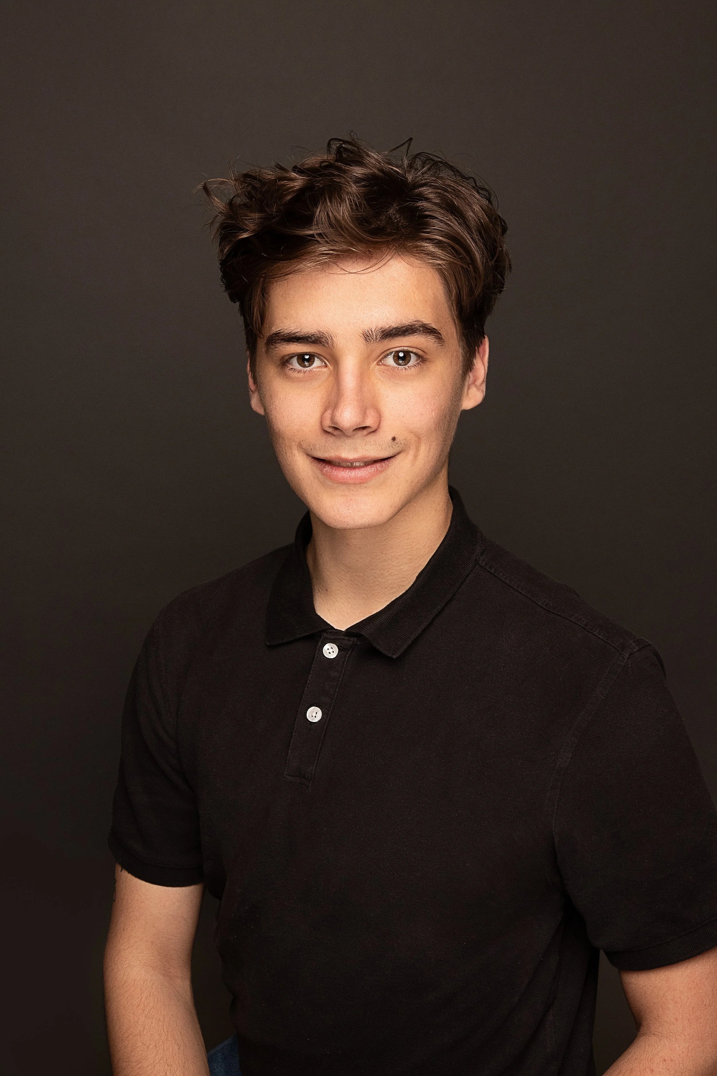 Portrait of a young man with light skin, short brown hair, wearing a black collared shirt, smiling softly, against a dark background.