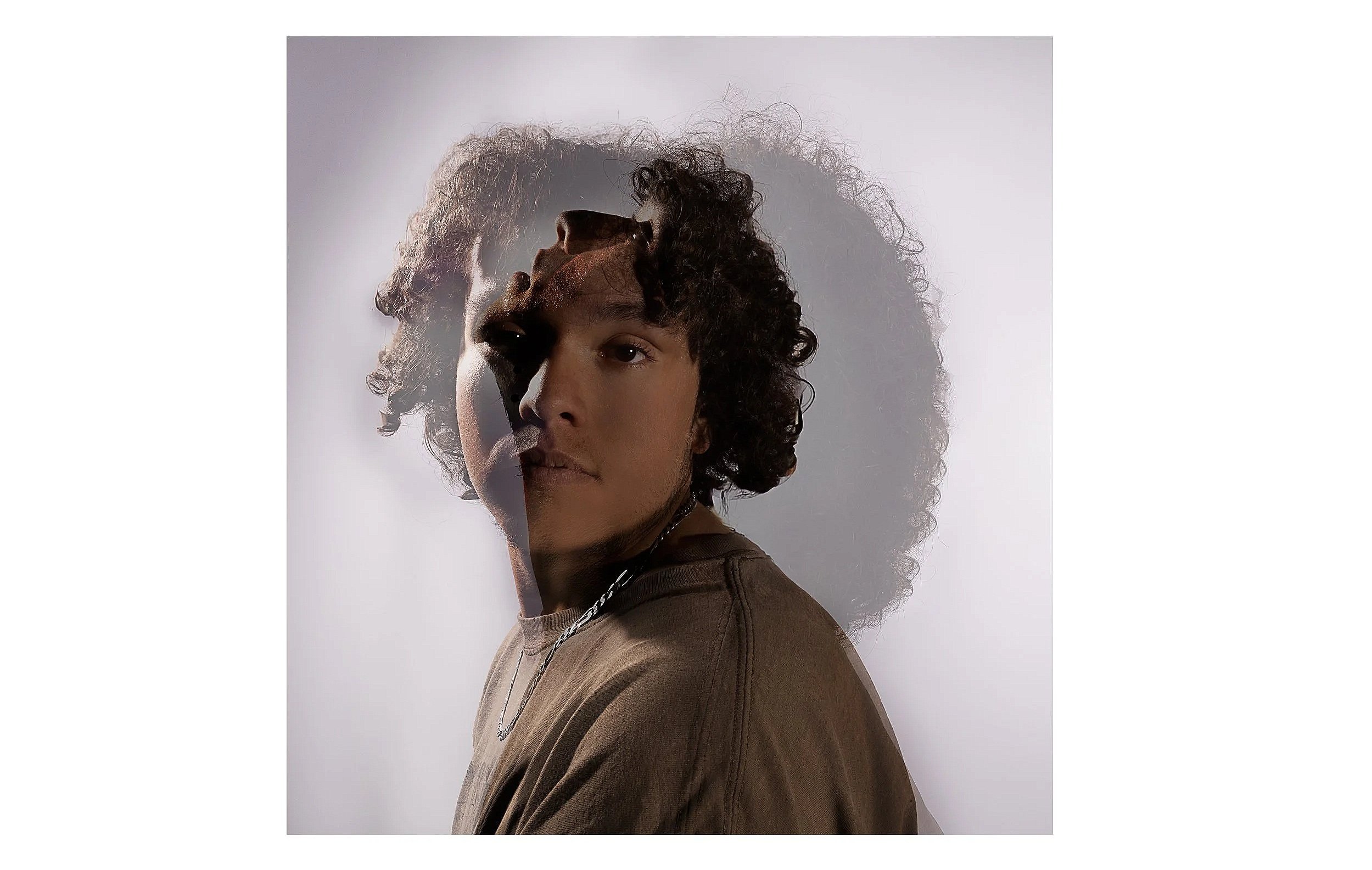 A person with short curly hair wearing a brown top and a silver chain necklace, with a double exposure effect creating a ghostly overlay, against a plain white background.