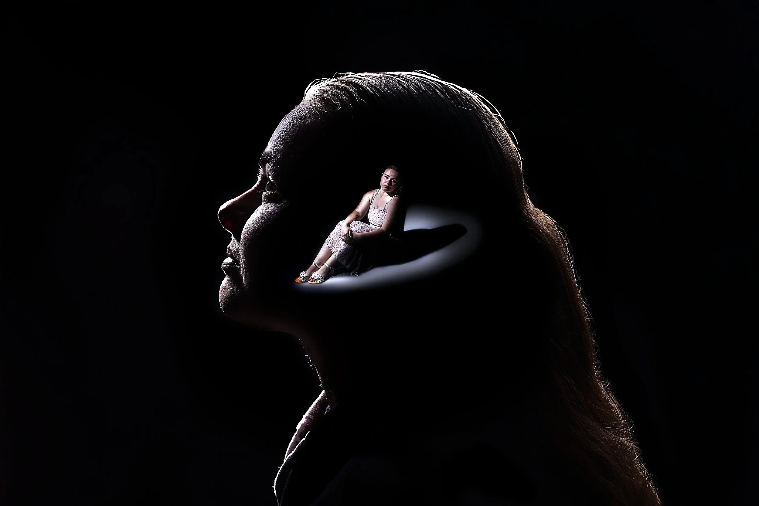 Silhouette of a woman's head in profile with a small woman sitting on her hand, set against a dark background, creating a surreal and artistic visual.