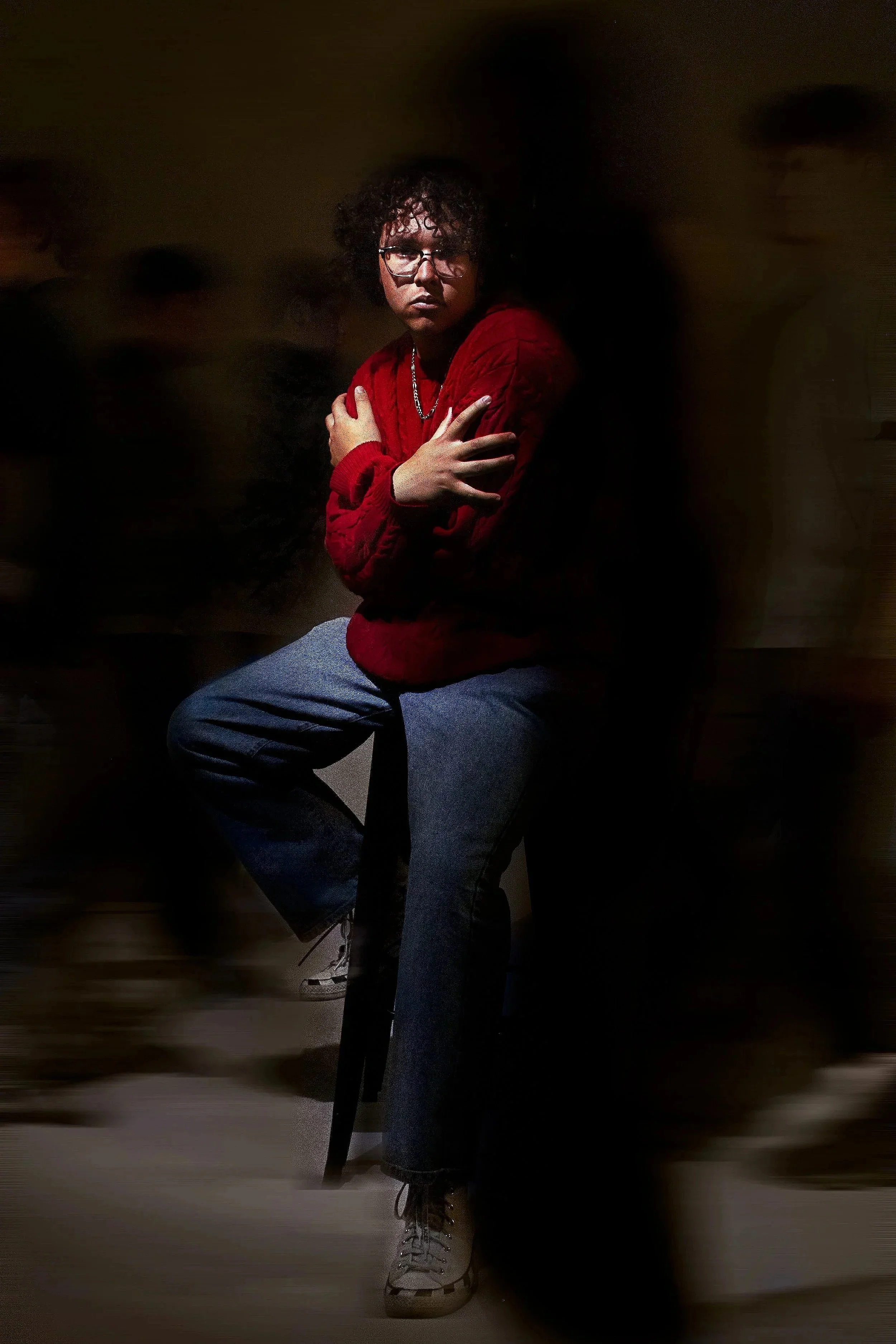 A young man with curly hair and glasses sitting on a black stool, wearing a red sweater and dark jeans, crossing his arms, in a dimly lit room with a dark background.