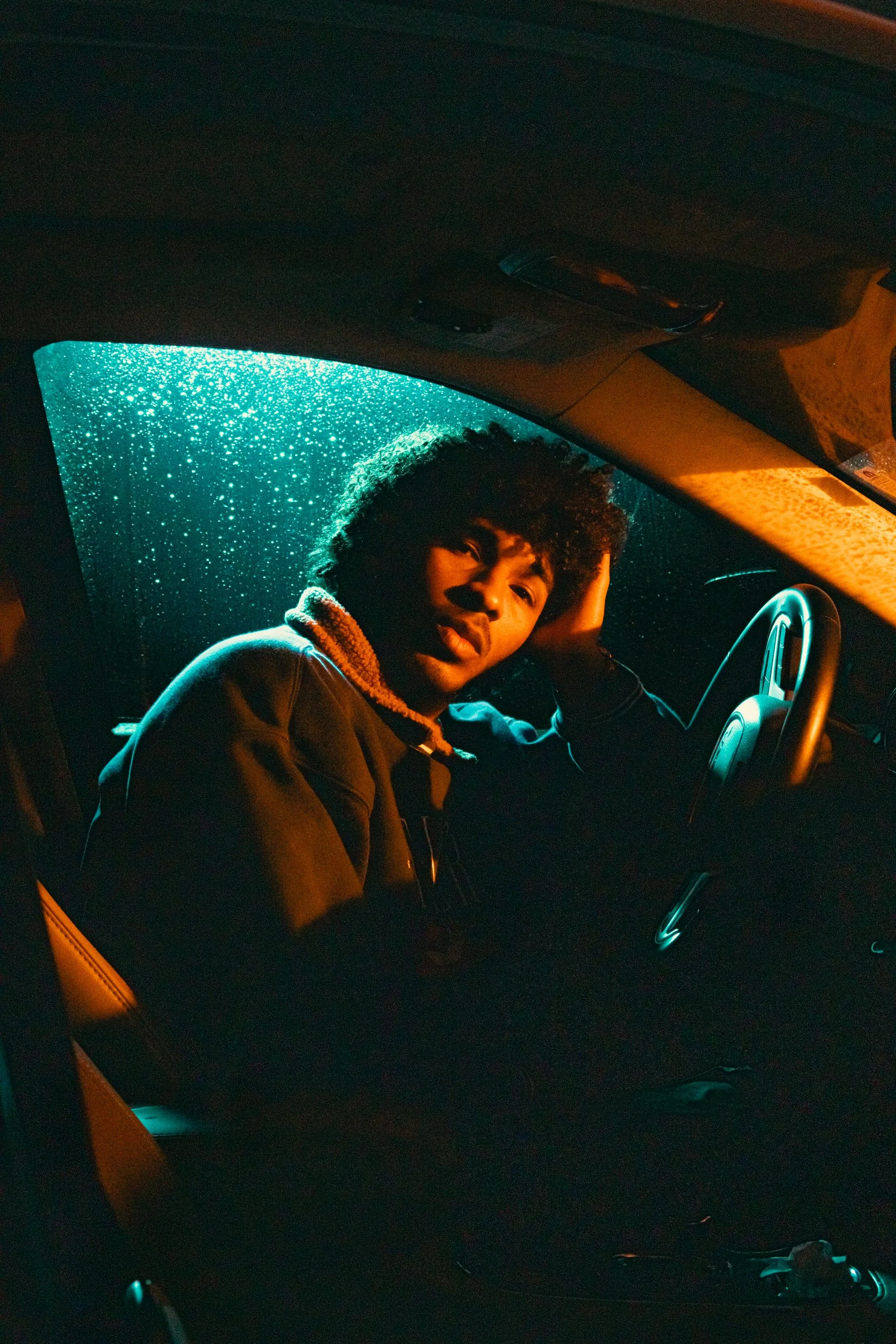 A young man with curly hair sitting inside a car at night, looking at the camera with a relaxed expression, rain visible on the window behind him, illuminated by a warm interior light.