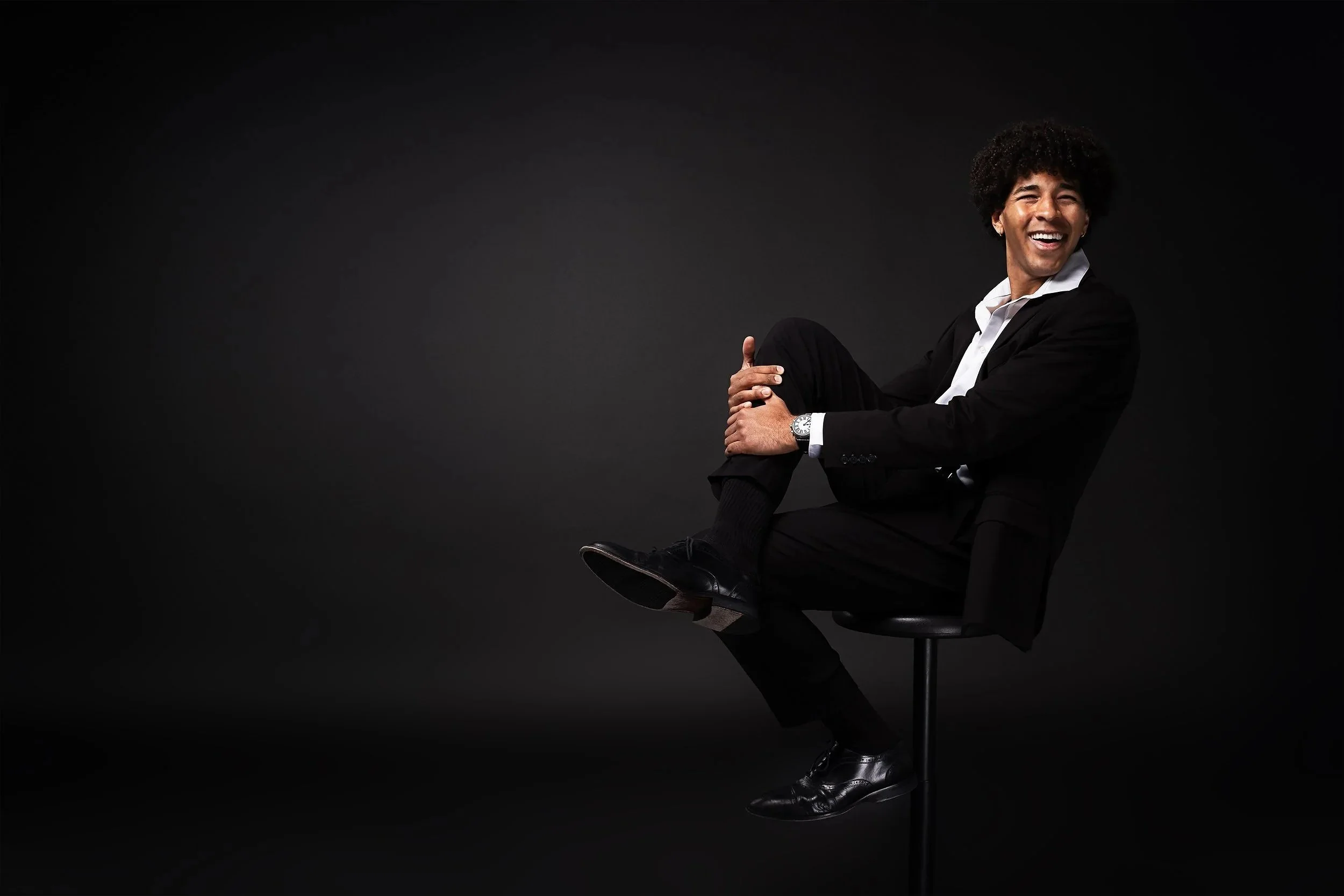 A man with curly hair and a bright smile, dressed in a black suit and white shirt, sitting sideways on a black stool against a dark background.