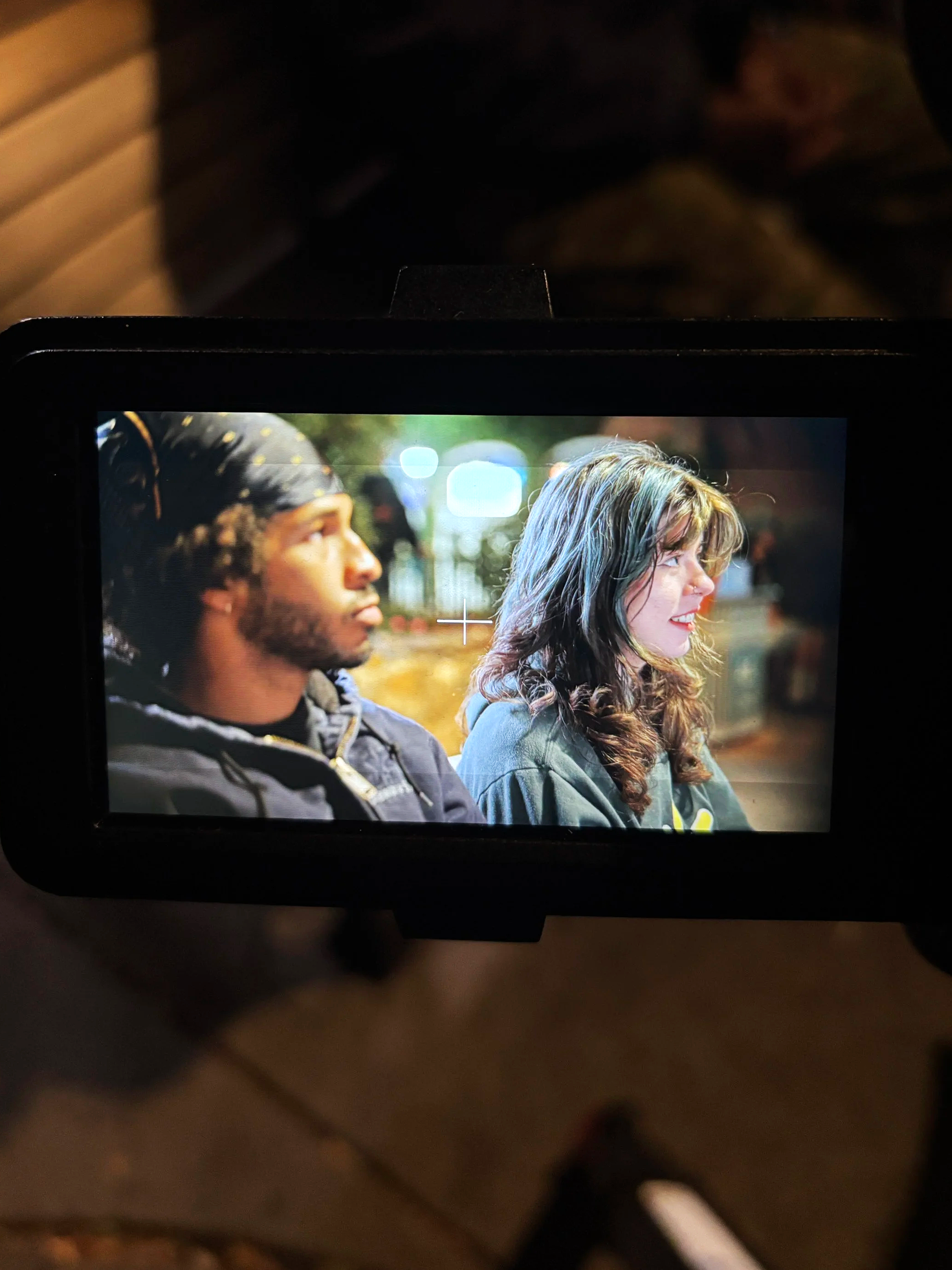 A camera screen showing a side profile of a young man with dark hair (Jordan Brumskill) and a young woman with long, wavy hair, both sitting outdoors - representing a stand-by moment on a Film Set.