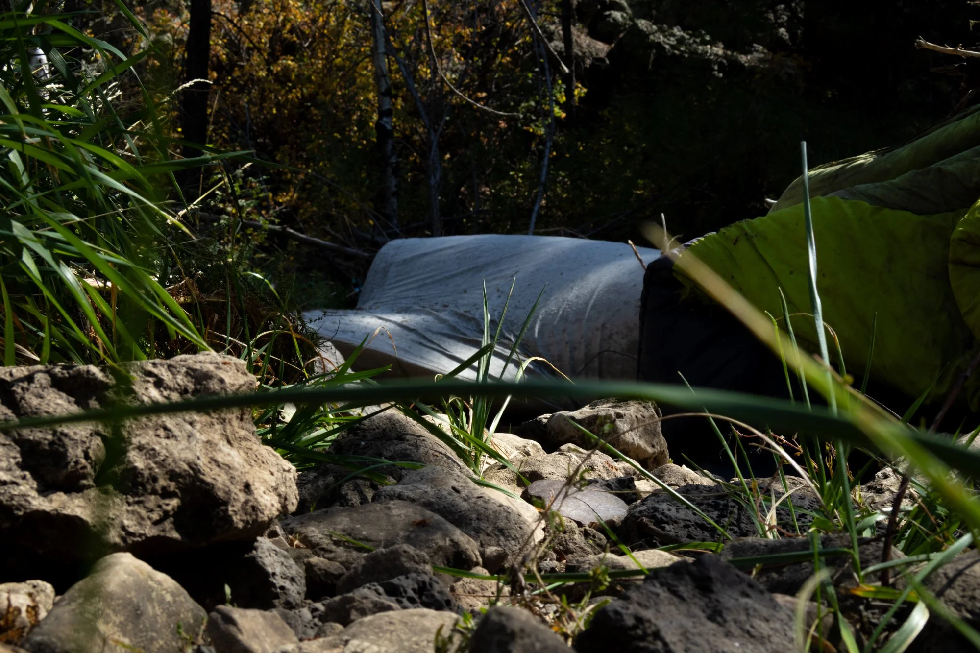 A person lying on the ground in a wooded area, covered with a green jacket, with the head obscured. The person is surrounded by rocks, dirt, and tall grass, with trees and foliage in the background.