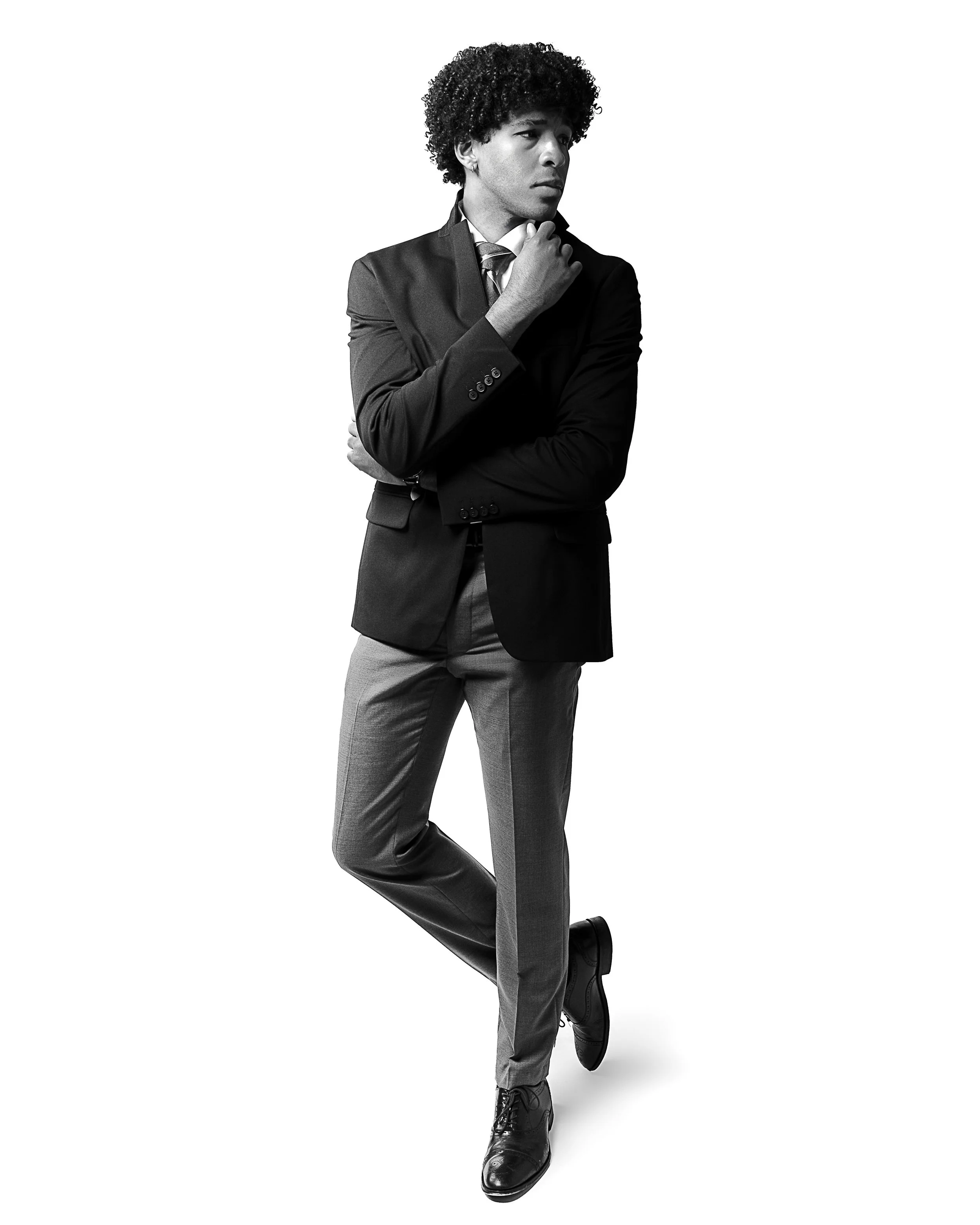 Black and white portrait of an African American man in a business suit, looking thoughtful with his hand on his chin, standing with one leg crossed over the other against a plain white background.