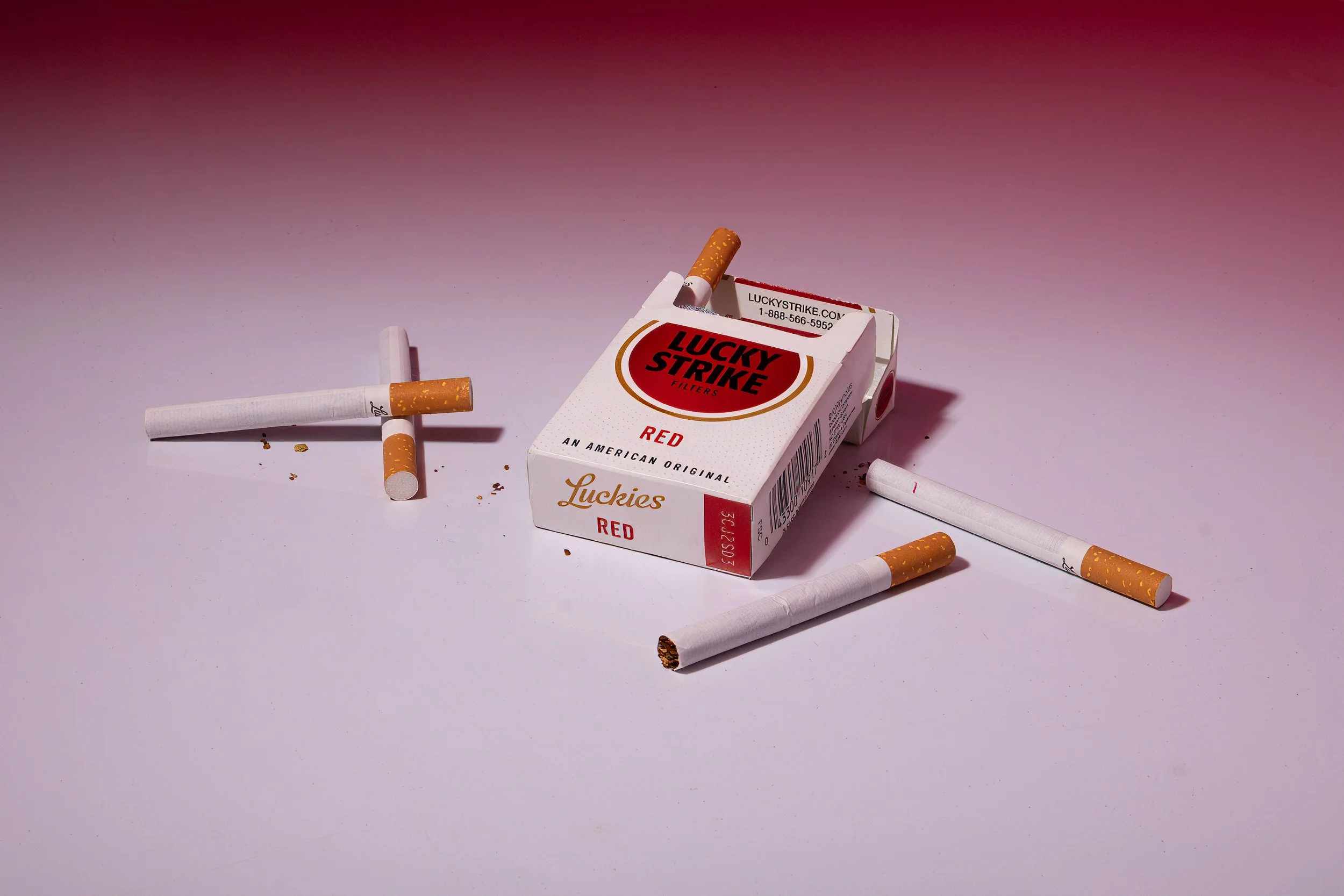 A box of Lucky Strike cigarettes with some cigarette butts and smoked cigarettes scattered around on a pink background.