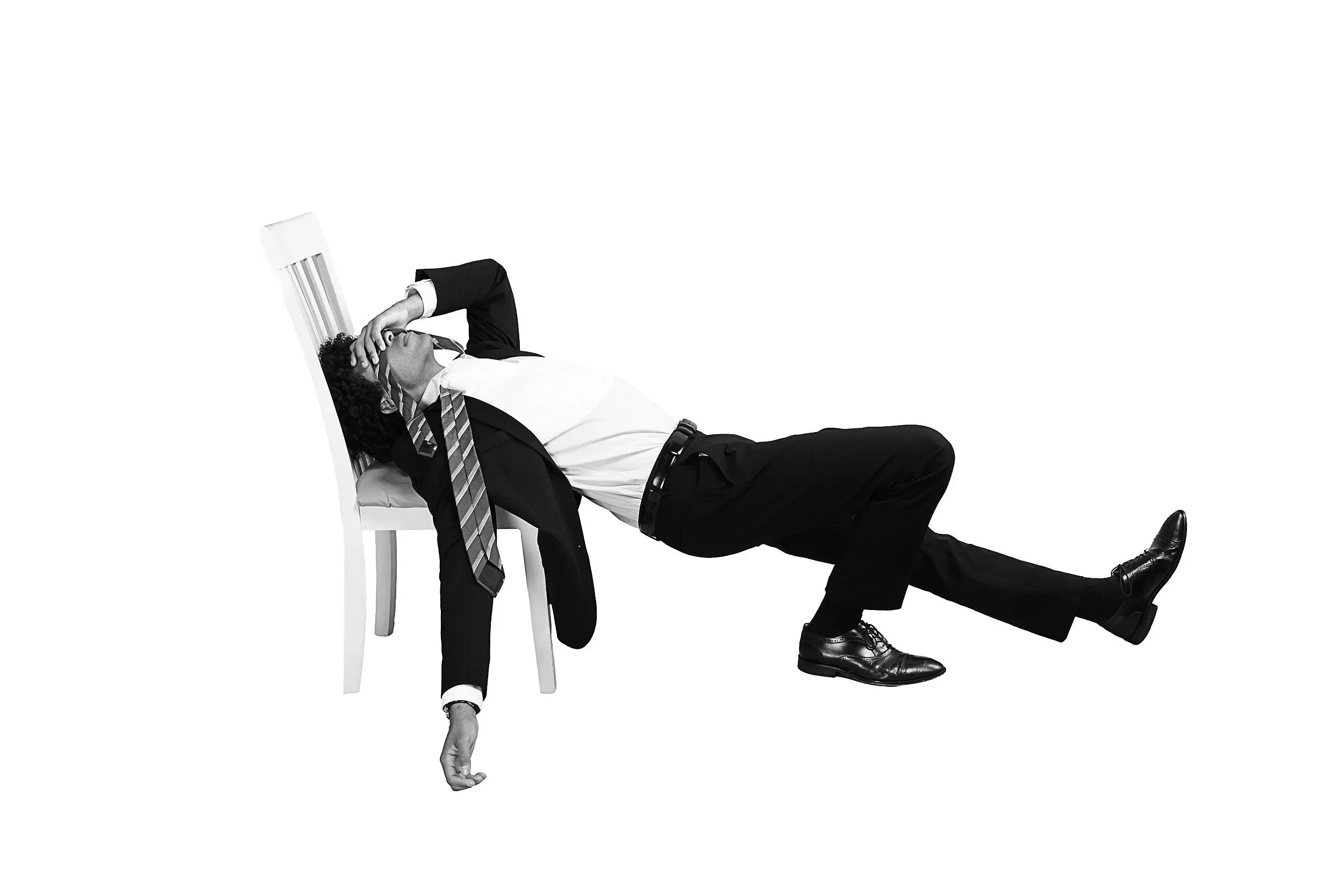A man in a business suit lying on the floor with one hand on his face and the other hanging down, and one leg bent, against a blank white background.
