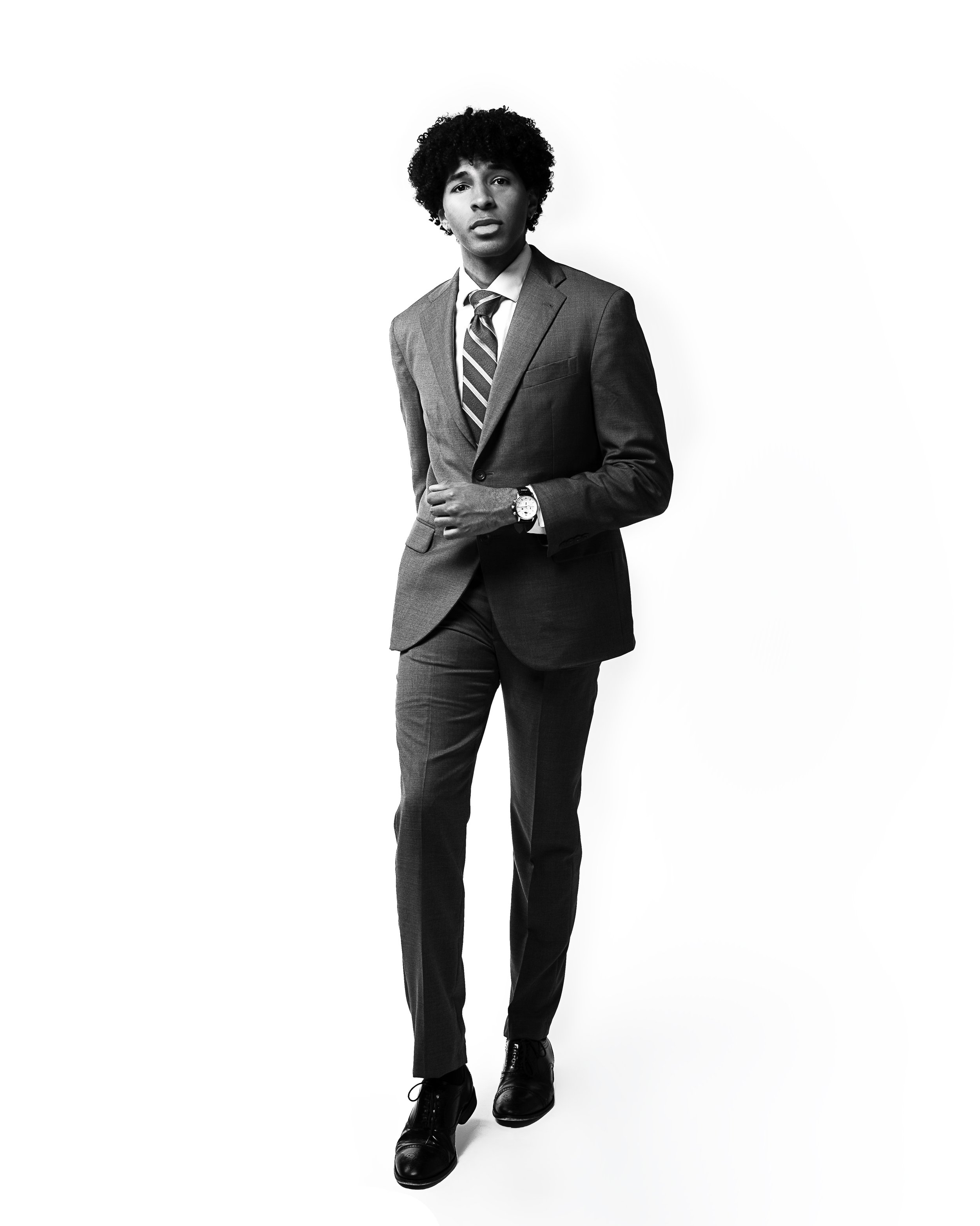 A young man with curly hair in a suit and tie, wearing black dress shoes and a wristwatch, standing against a plain white background.