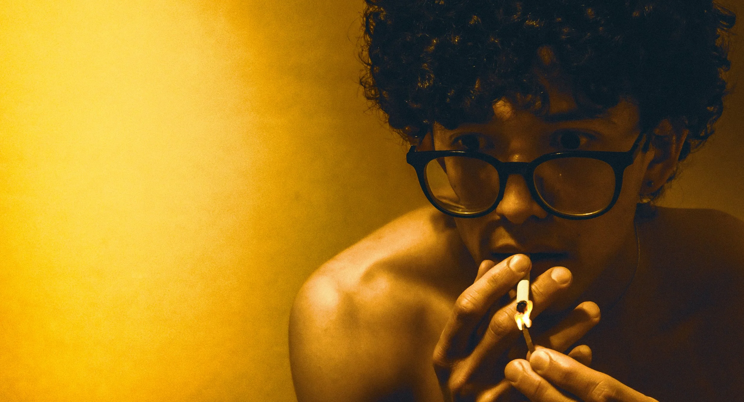 A person with curly hair and glasses smoking a cigarette against a yellow background.