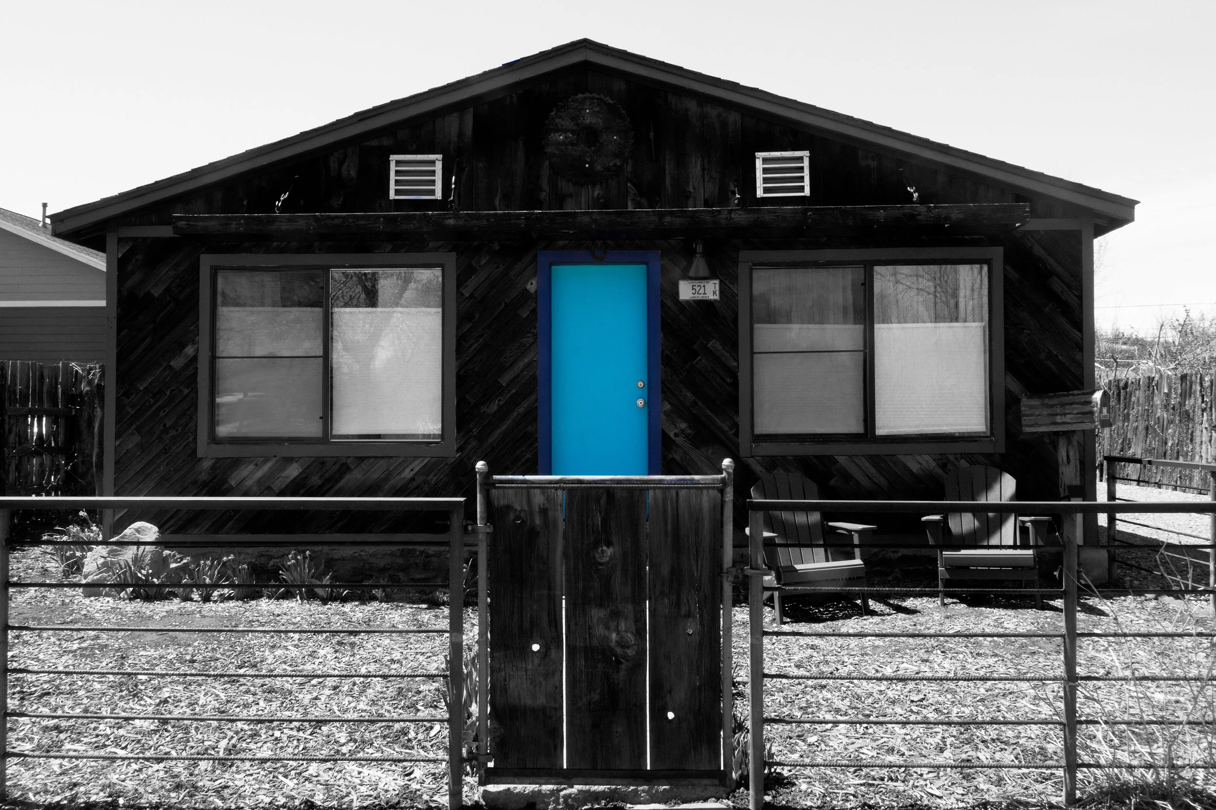 A black-and-white photo of a house with a bright blue door, two large front windows, and a small front yard with a wooden fence and a swing set.
